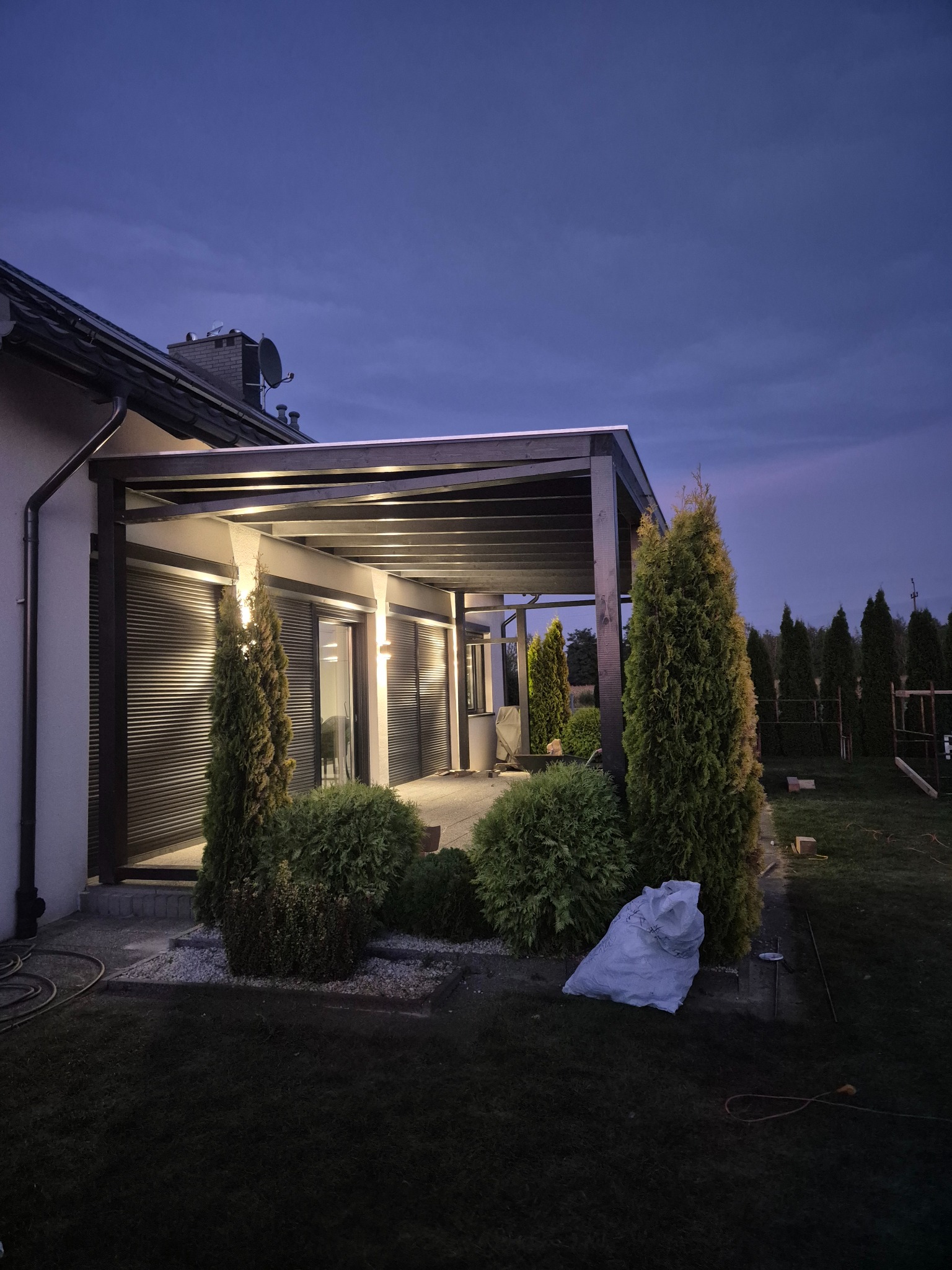 Nowoczesna pergola z roletami zewnętrznymi i oświetleniem, otoczona zielenią w wieczornym świetle. Widoczny worek z odpadami po pracach.