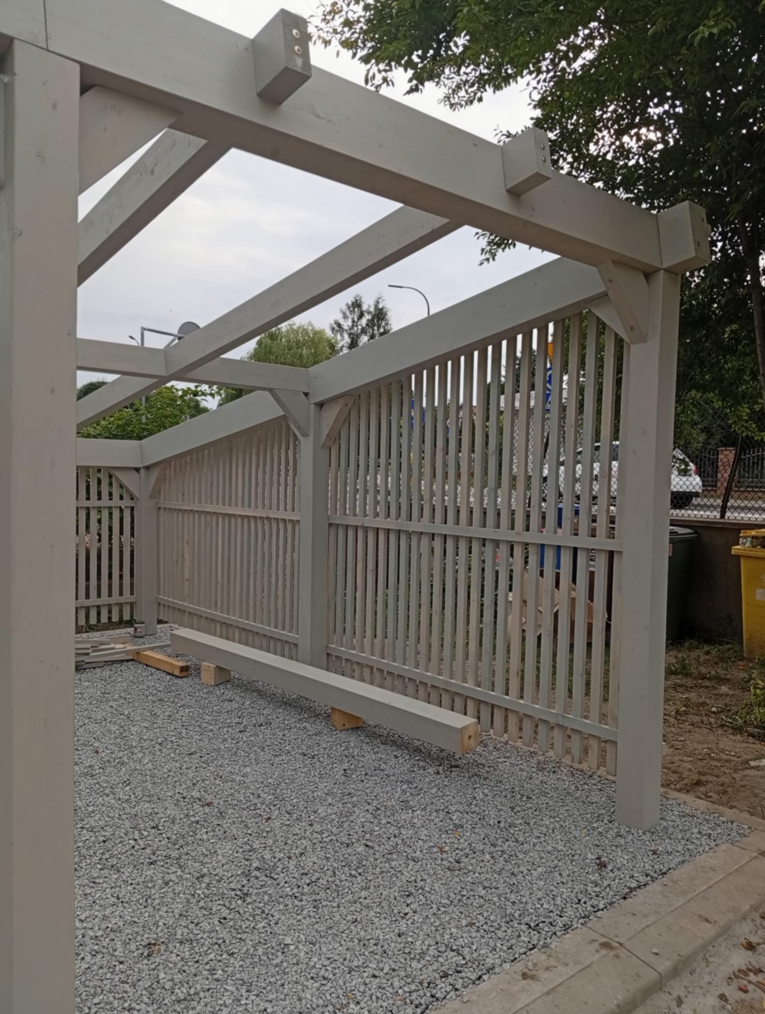 Szara drewniana pergola z pionowymi listwami, częściowo zmontowana, na żwirowym podłożu. Widoczne elementy konstrukcyjne i fragmenty otoczenia.