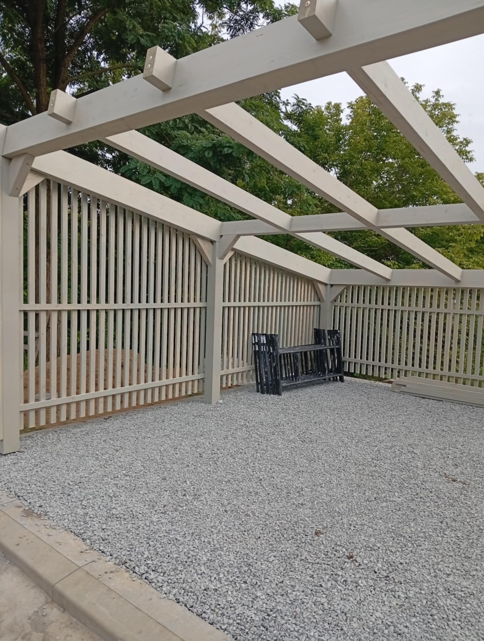 Drewniana pergola z ażurowymi ścianami, pomalowana na jasny kolor, z podłożem wysypanym drobnym kamieniem. W tle widoczne drzewa.