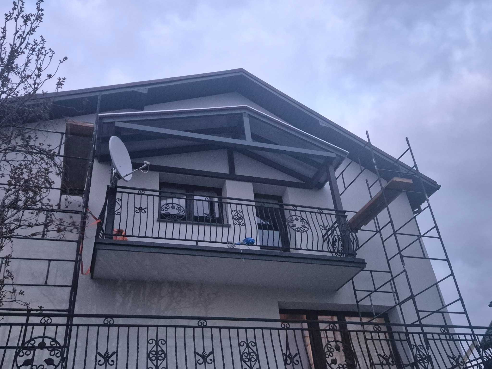 Dwupiętrowy dom z balkonem w trakcie prac wykończeniowych. Widoczne rusztowania i elementy elewacji. Ujęcie z dołu, pochmurne niebo w tle.