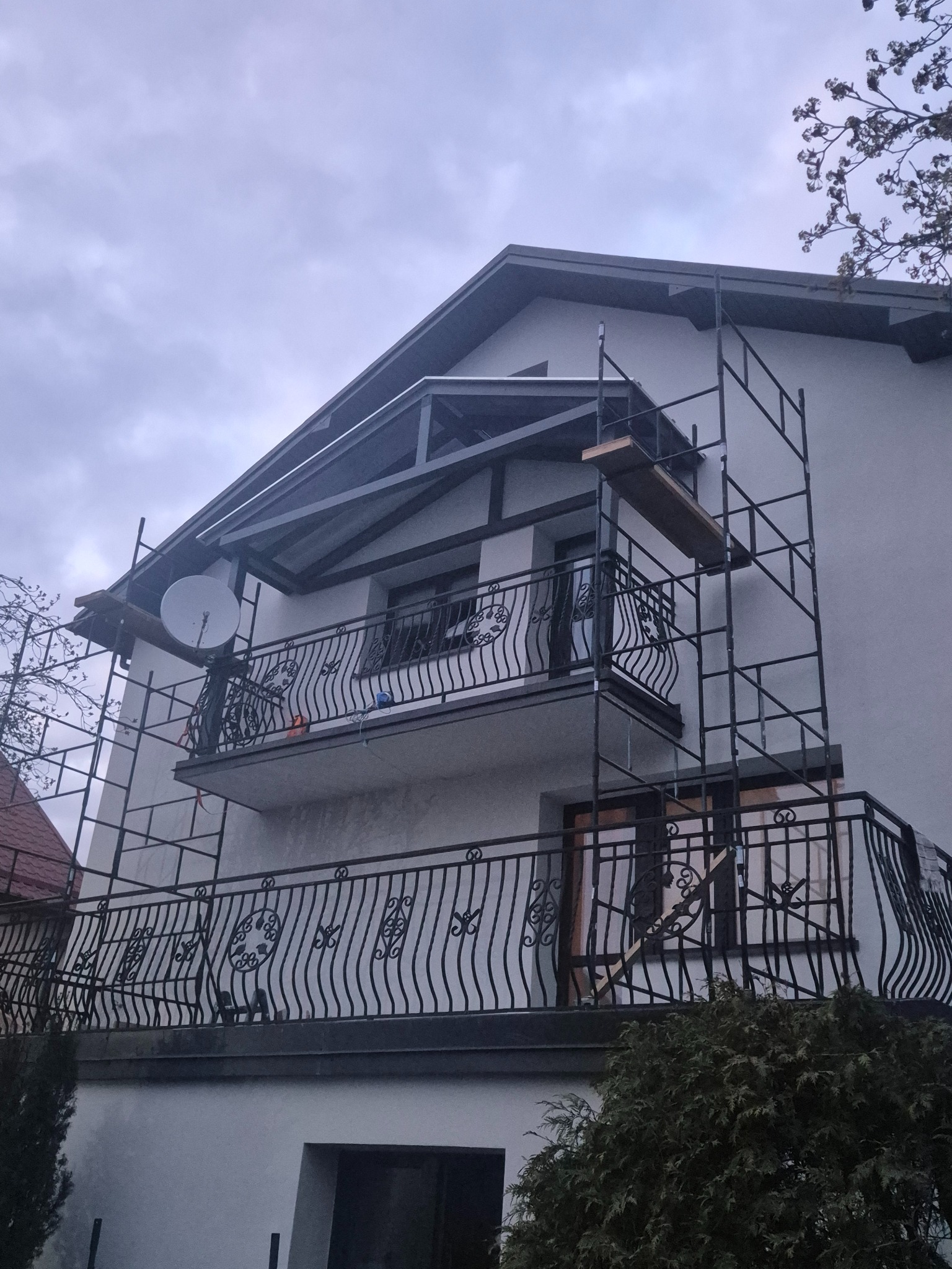 Elewacja domu w trakcie prac z rusztowaniem. Widoczny balkon z ozdobną balustradą i satelita. Szare niebo w tle.
