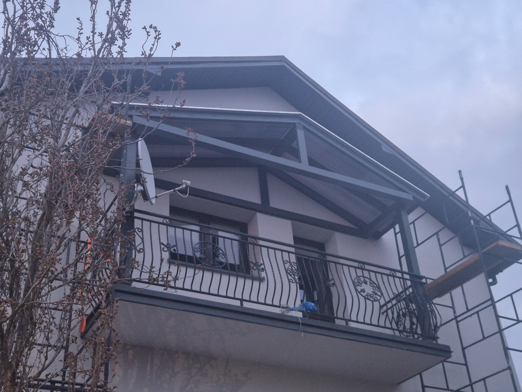 Elewacja domu z balkonem i metalową balustradą, widoczne rusztowanie. Drzewo z pąkami po lewej stronie. Ujęcie z dołu, pochmurne niebo w tle.