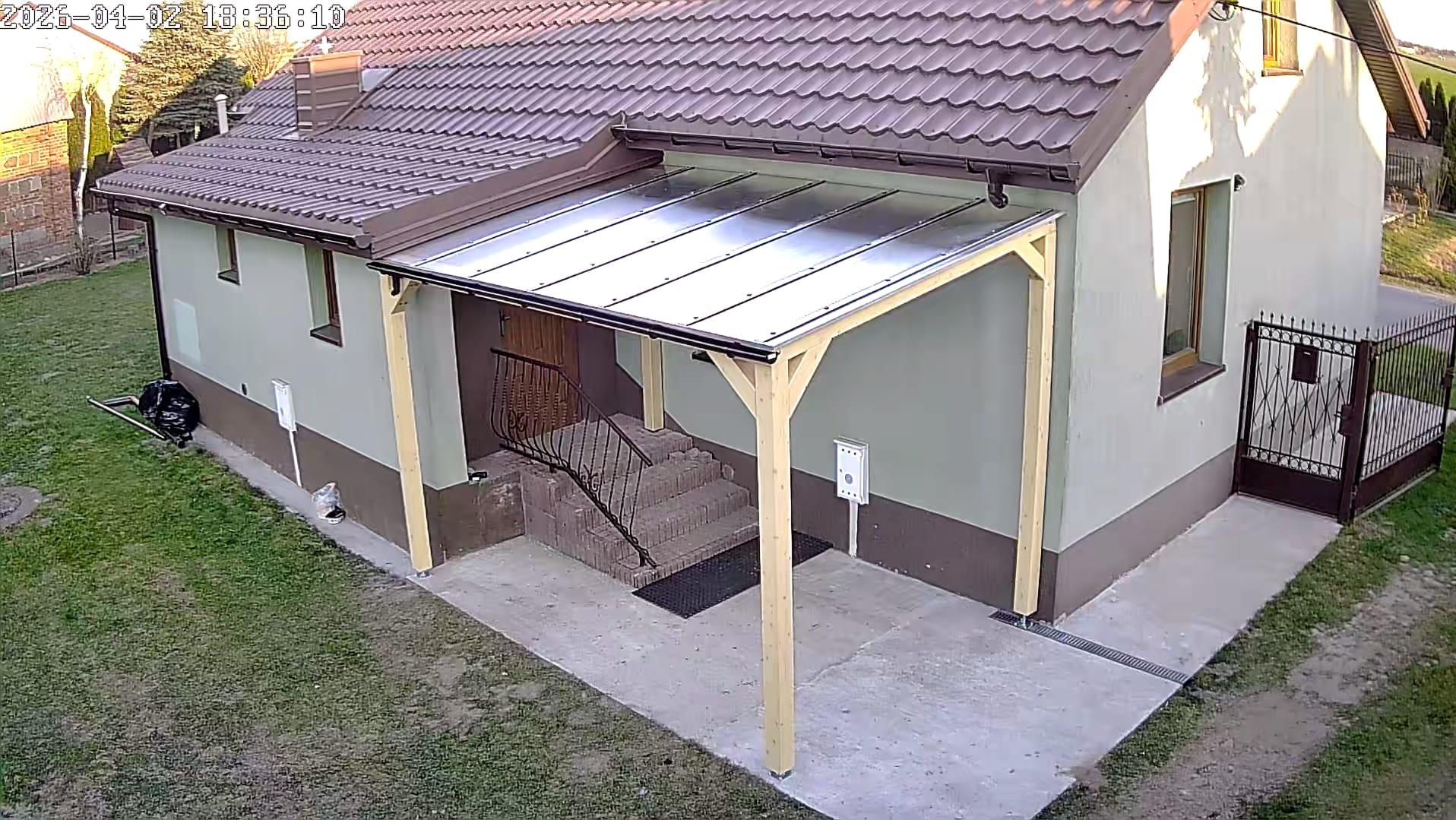 Zadaszenie nad wejściem do domu z drewnianą konstrukcją i przezroczystym pokryciem, widok z góry, brama w tle.