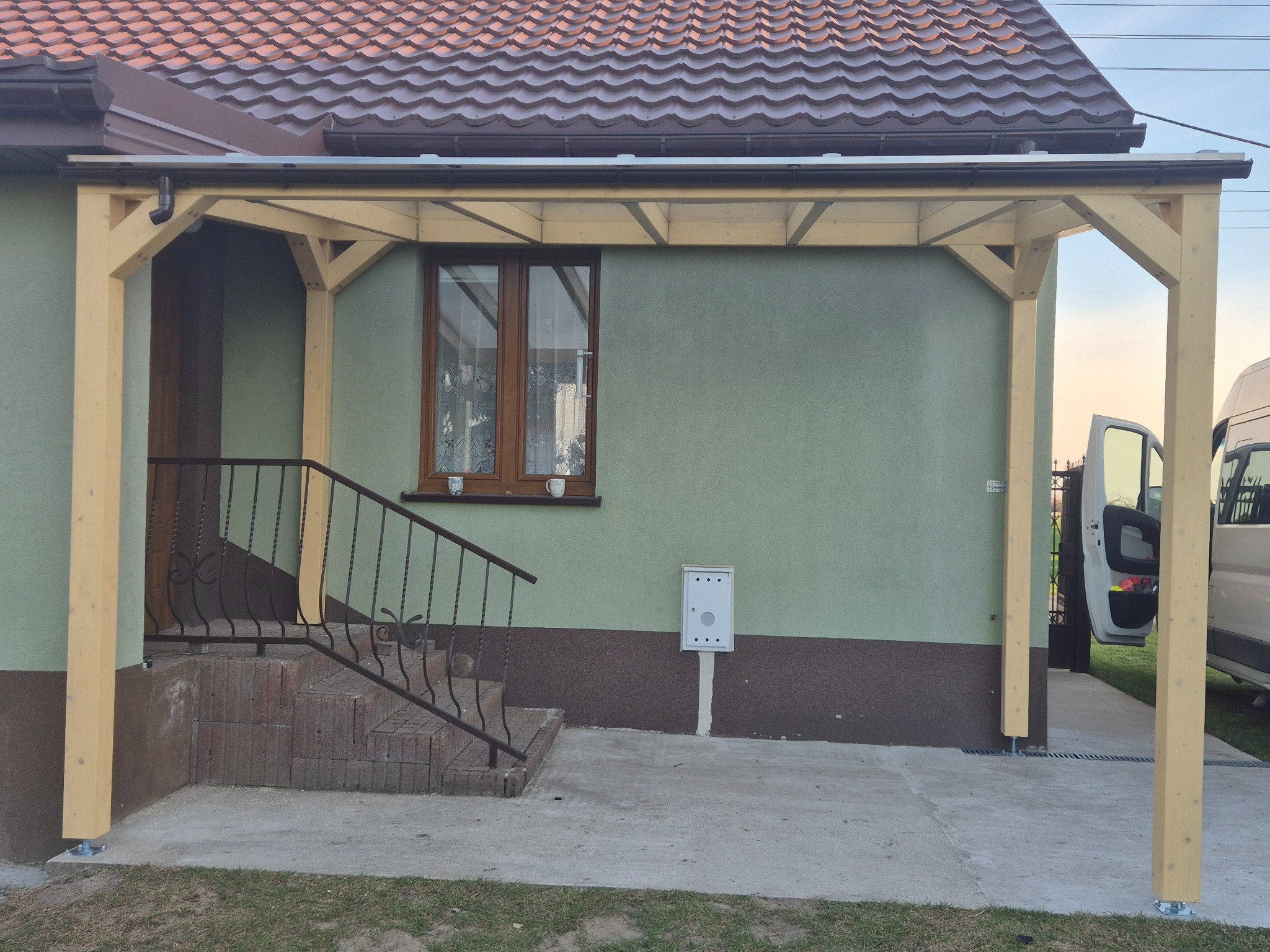 Zadaszenie wejścia do domu z jasnego drewna, widok z zewnątrz. Elementy konstrukcyjne złączone śrubami. W tle ściana domu z oknem i skrzynką pocztową.