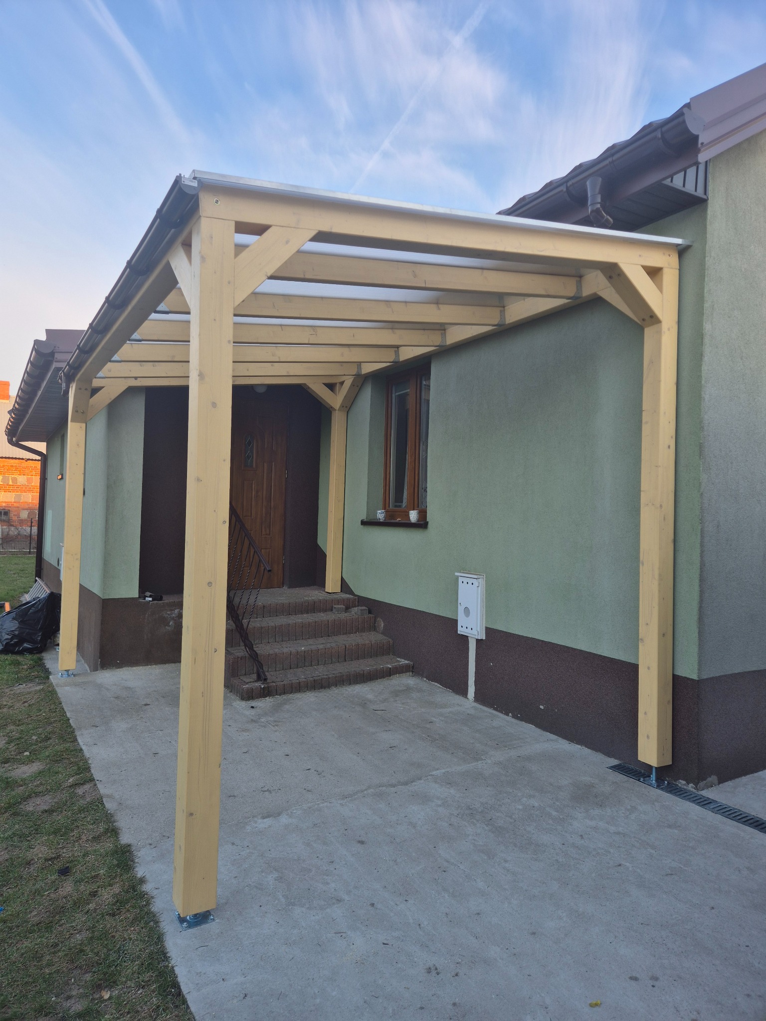 Drewniana pergola z przezroczystym zadaszeniem przy domu z zieloną elewacją. Widoczne schody wejściowe i skrzynka pocztowa. Ujęcie z dołu, niebieskie niebo.