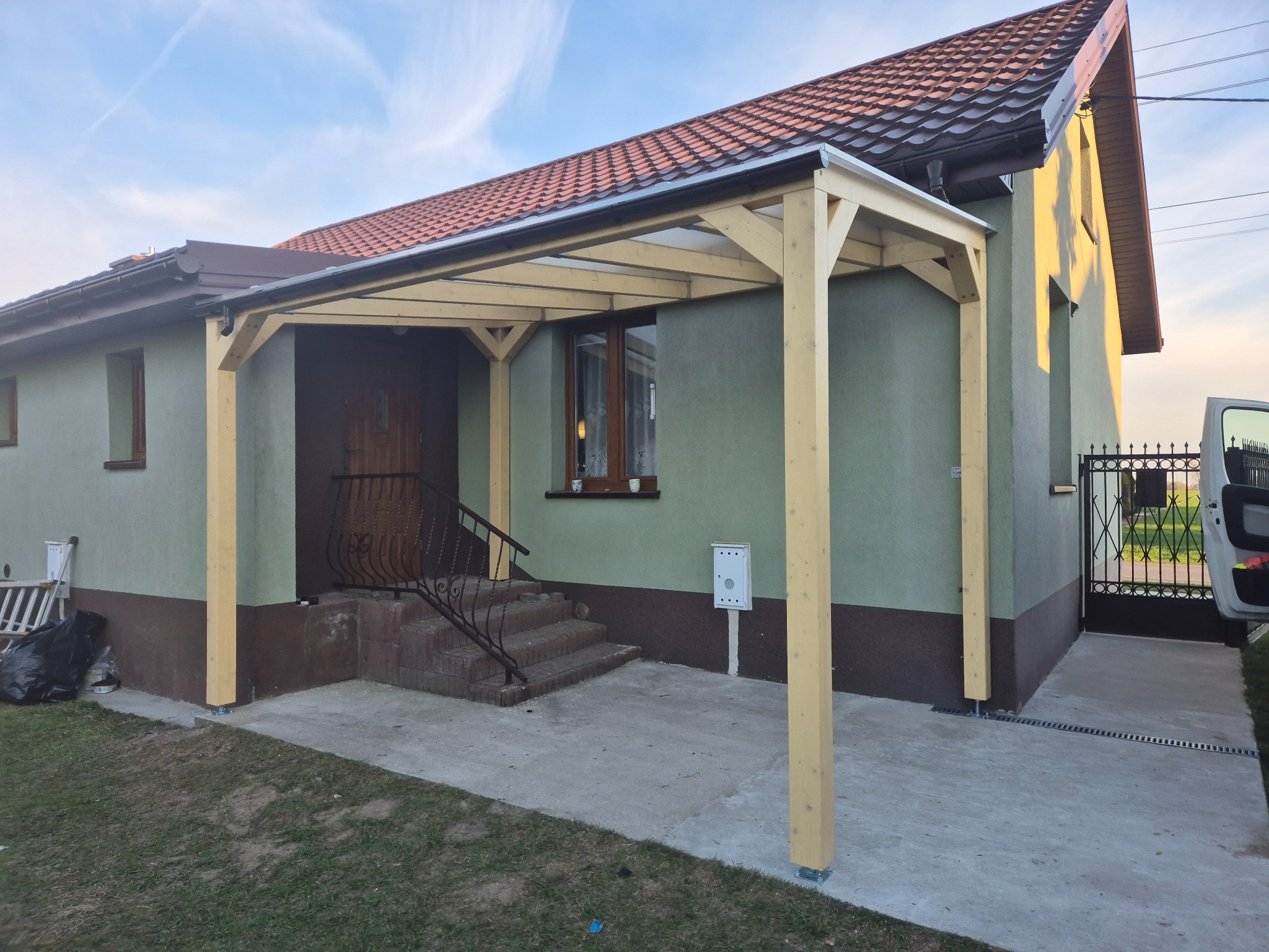 Zadaszenie nad wejściem do domu w Mszczonowie. Drewniana konstrukcja z przezroczystym dachem, widoczne schody i elewacja w odcieniach zieleni i brązu. Detal architektoniczny.