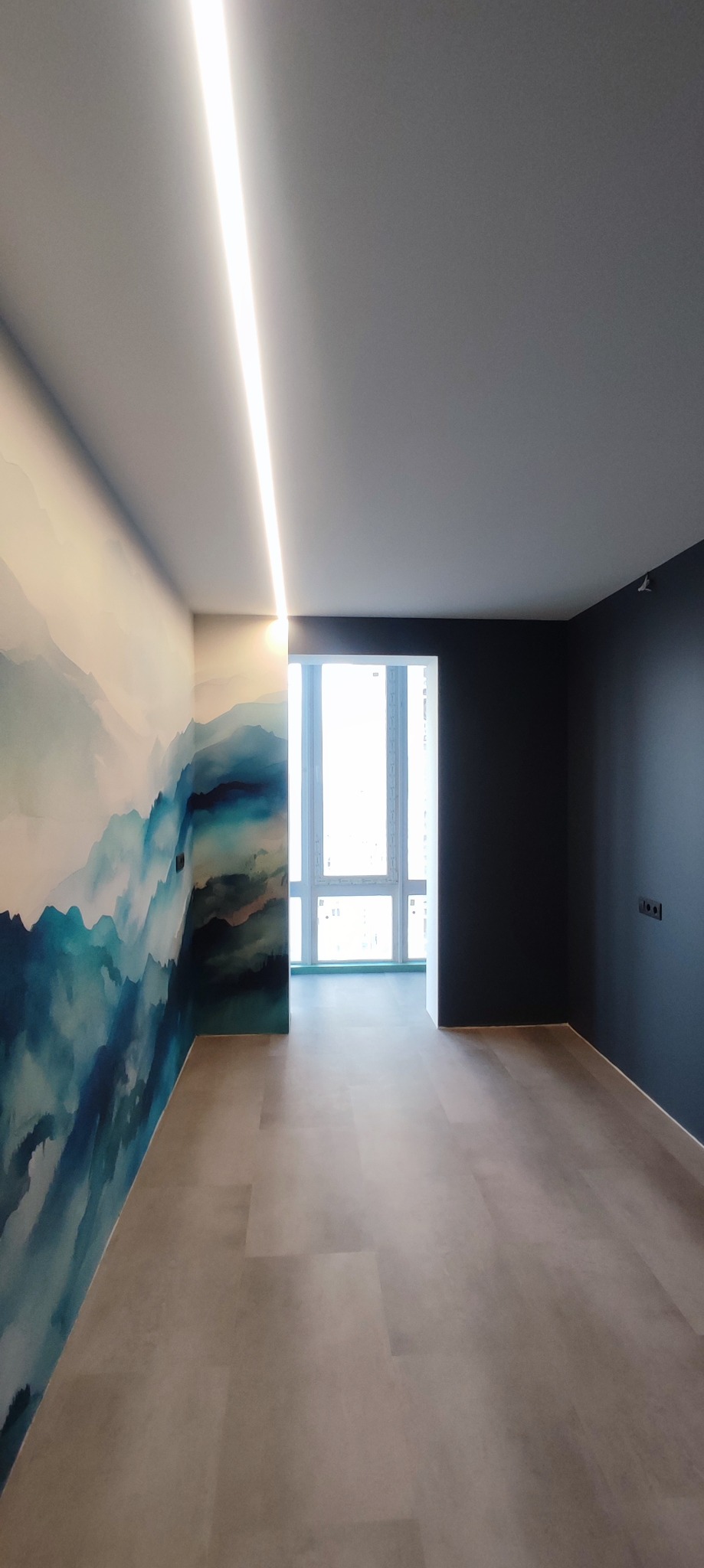 Wnętrze pustego pokoju z podświetleniem LED w suficie, ściana z wzorem górskim w odcieniach błękitu i granatu, oraz ciemnoszara ściana z gniazdkiem elektrycznym.
