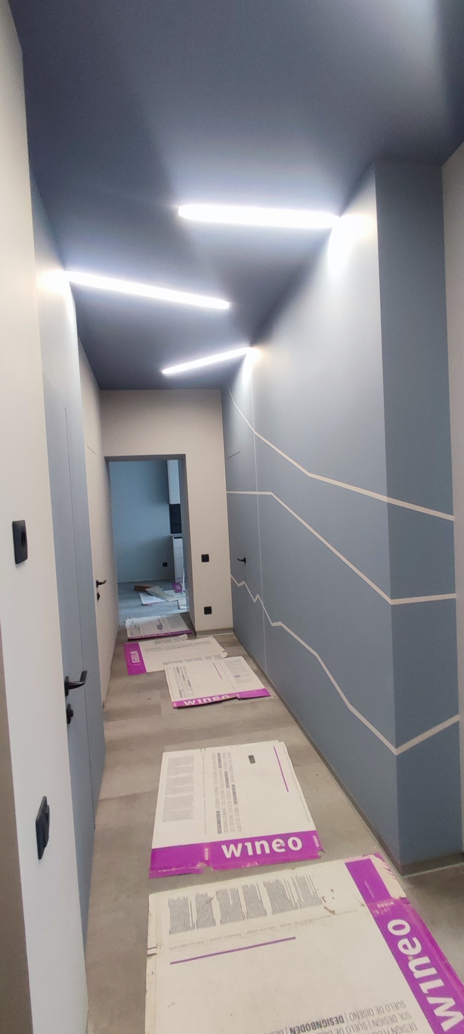 Korytarz w trakcie remontu, ściany pomalowane na szaro-niebieski kolor z białymi geometrycznymi wzorami, podłoga zabezpieczona kartonami firmy Wineo, sufit pomalowany na ciemniejszy odcień szarości...