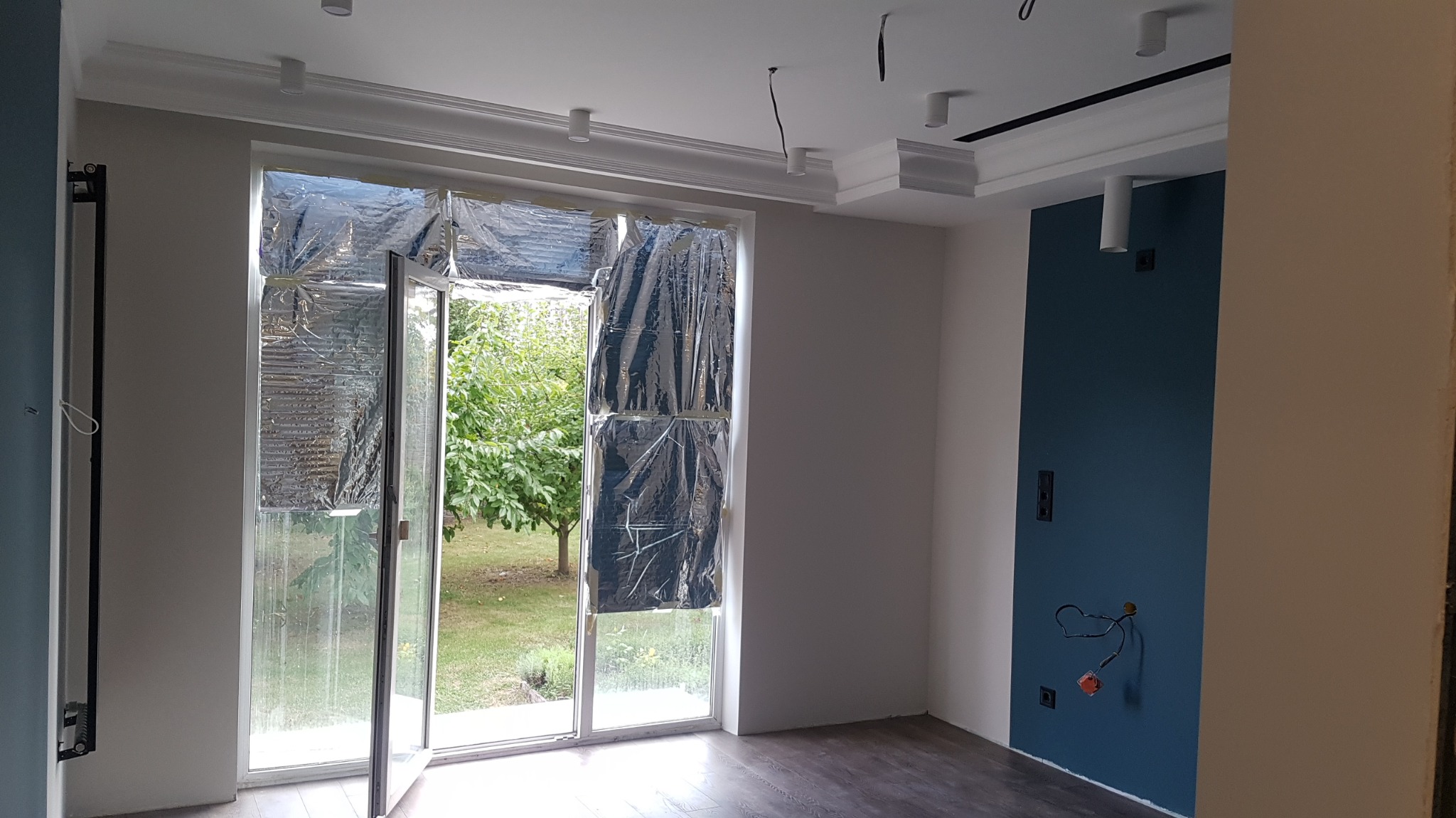 Wnętrze pokoju w trakcie remontu: okno zabezpieczone folią ochronną, ściana pomalowana na dwa kolory, widoczne wystające kable elektryczne.
