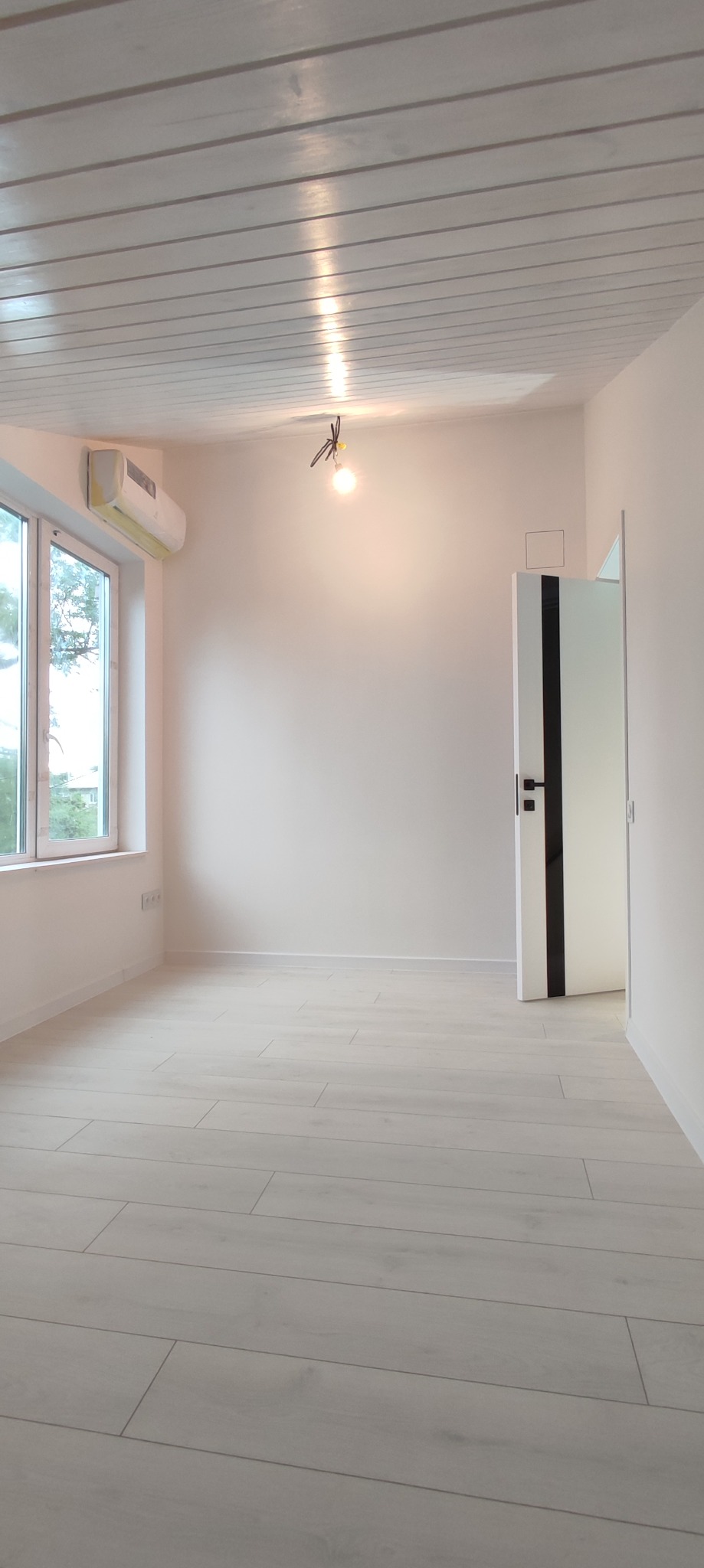 Jasne, puste pomieszczenie po remoncie z drewnianą podłogą, białymi ścianami i sufitem z paneli, oświetlone pojedynczą żarówką.