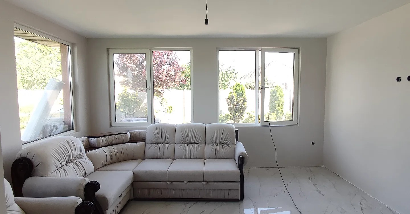 Jasne wnętrze po świeżym malowaniu ścian na biało, widoczne gniazdka elektryczne bez osłon, marmurowa podłoga i narożna sofa w odcieniach beżu.