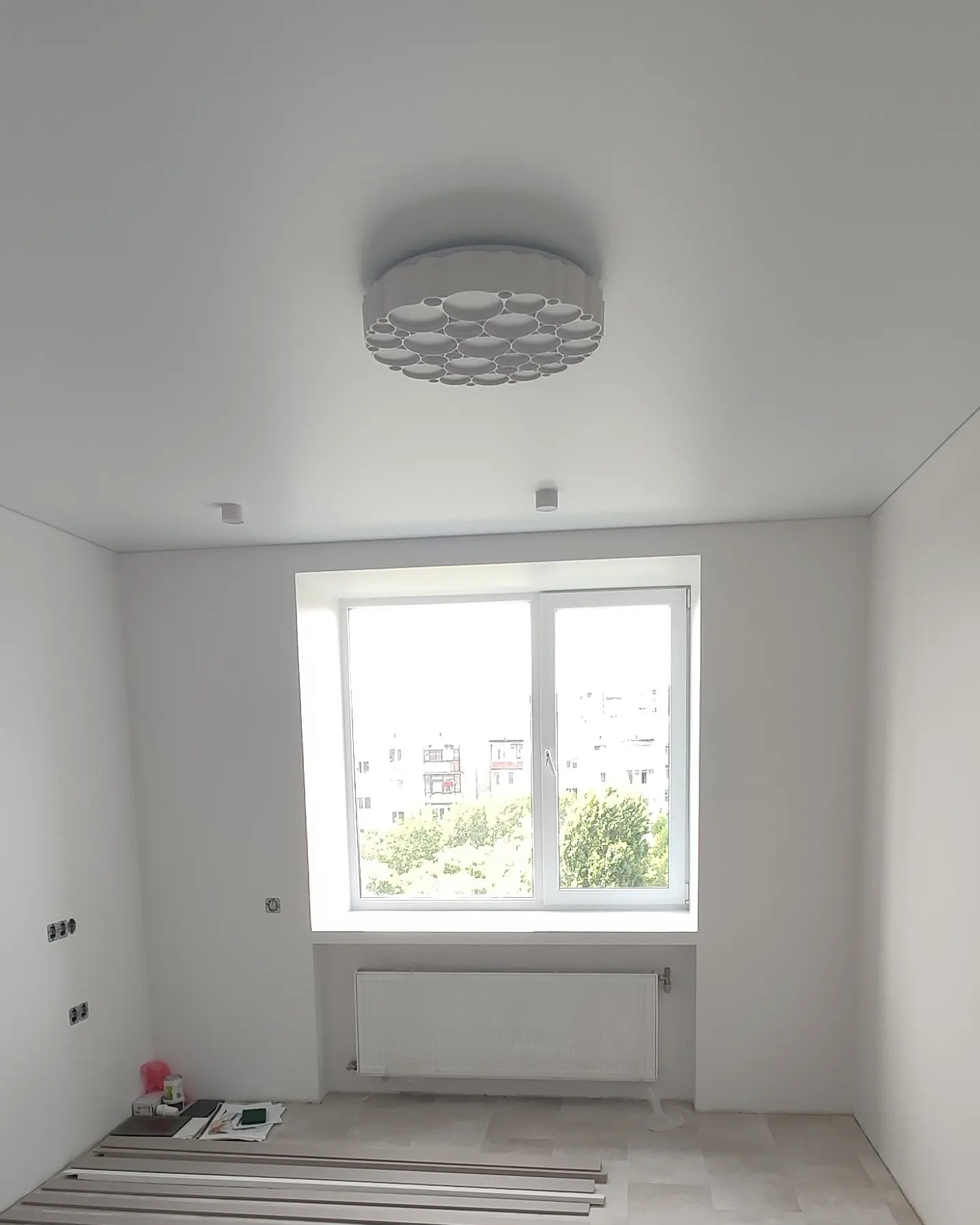 Wnętrze pokoju po świeżym malowaniu na biało, widoczne puszki elektryczne, grzejnik pod oknem i nowoczesna lampa sufitowa.