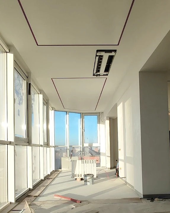 Wnętrze w trakcie malowania: biały sufit z zaznaczonymi bordowymi liniami, widoczne otwory wentylacyjne, duże okna z widokiem na miasto, grzejniki i narzędzia malarskie na podłodze.