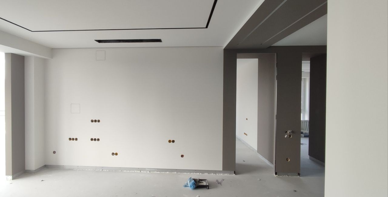 Wnętrze w trakcie przygotowania do malowania: ściany zagruntowane, widoczne puszki instalacyjne, listwy przypodłogowe zabezpieczone folią, na podłodze kuweta malarska.