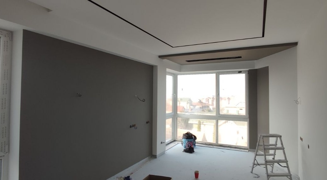 Pokój po częściowym malowaniu: szara ściana kontrastuje z białą, widoczne wystające kable elektryczne, drabina, okno z widokiem na zabudowę miejską.