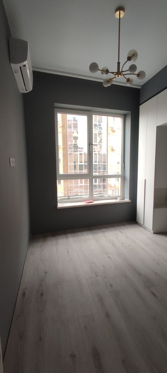 Pokój po remoncie: szare ściany, jasna podłoga, biała szafa, nowoczesna lampa sufitowa, widok z okna na budynek mieszkalny.