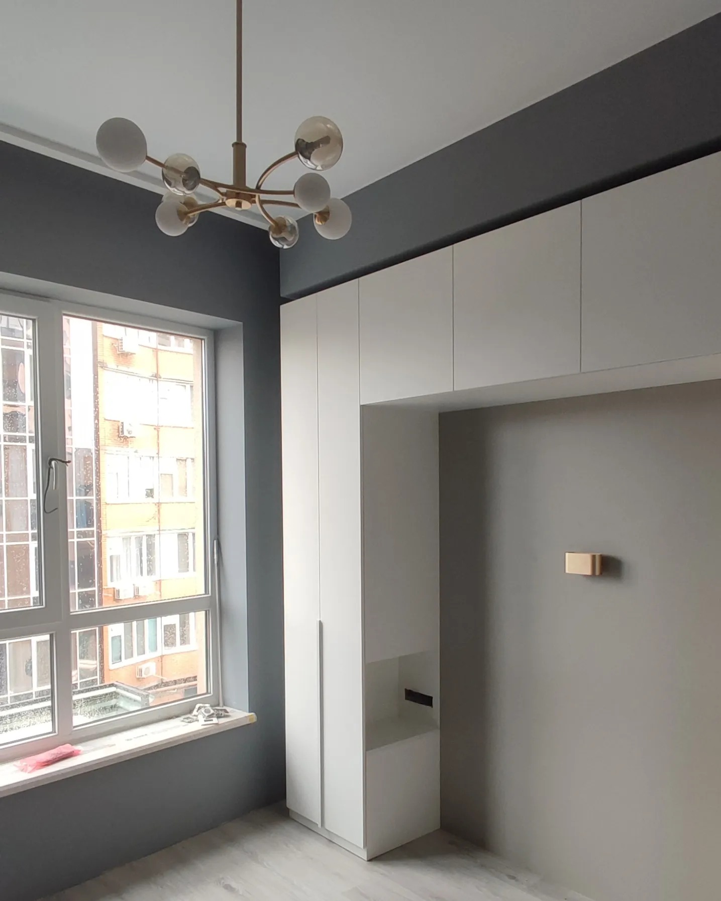 Pokój po remoncie: szara ściana, biała zabudowa meblowa, nowoczesny żyrandol z kulistymi kloszami, widok z okna na blok mieszkalny.