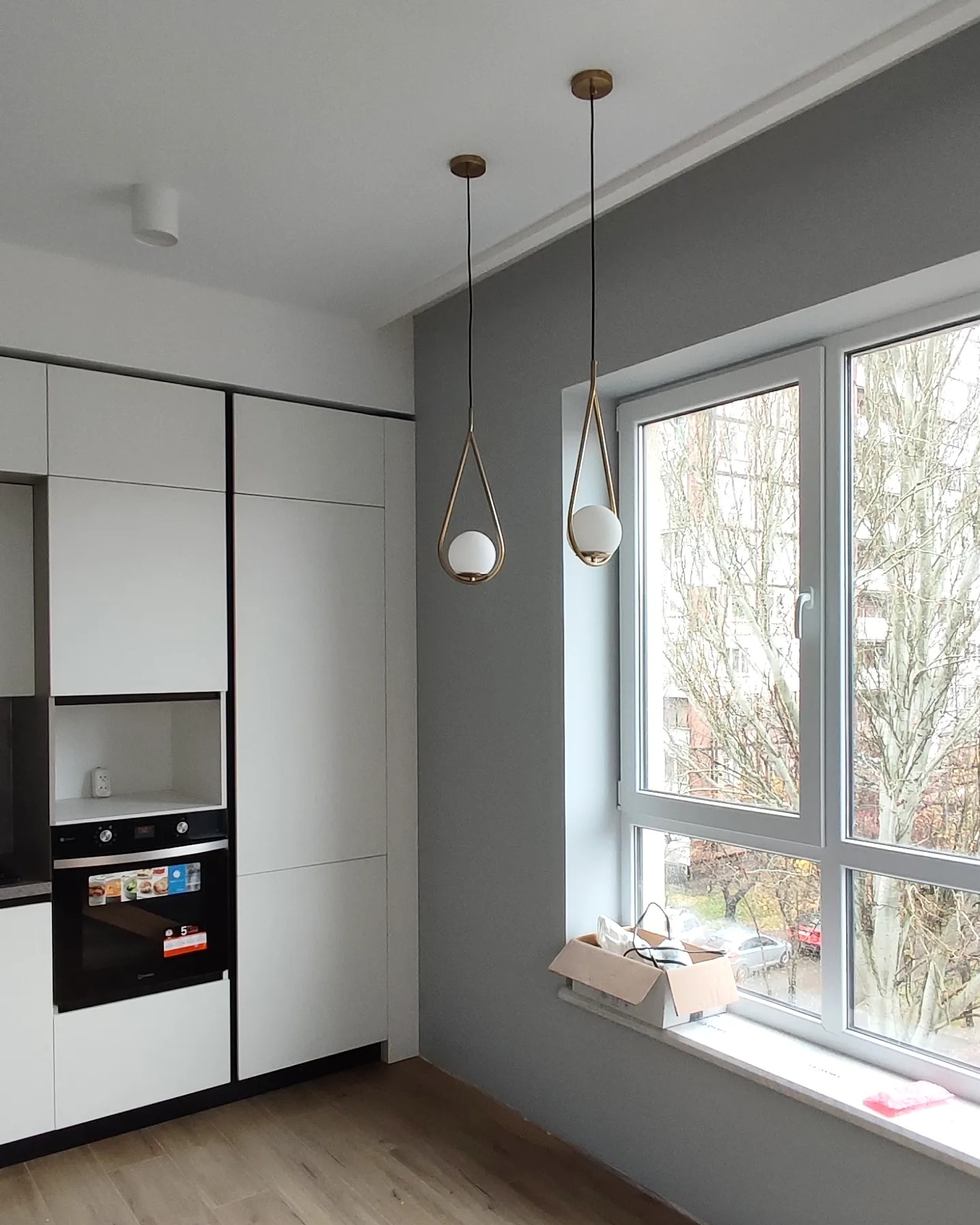 Szara ściana po malowaniu w nowym mieszkaniu, widoczne lampy wiszące z kulistymi kloszami, białe okno z widokiem na drzewa, karton na parapecie i biała zabudowa kuchenna.