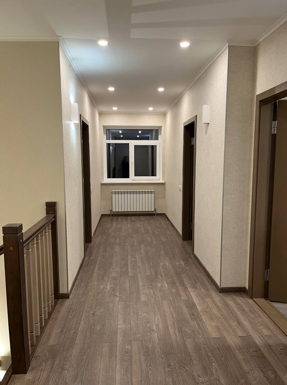 Wykończony korytarz z drewnopodobną podłogą, świeżo pomalowanymi ścianami w odcieniach beżu i białymi listwami przypodłogowymi, oświetlony punktowymi lampami sufitowymi.