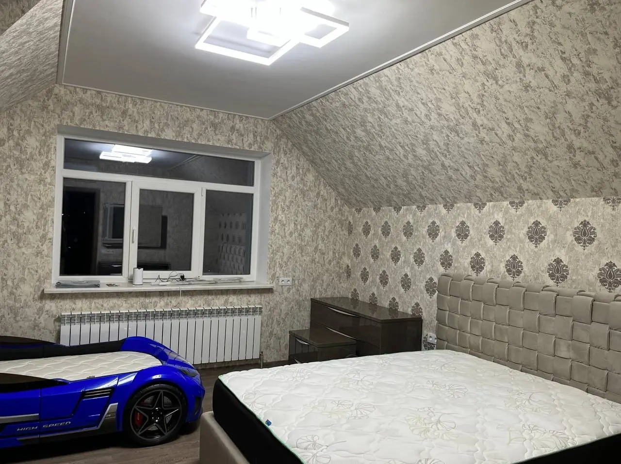 Pokój na poddaszu z tapetą w neutralnych odcieniach, łóżkiem w kształcie samochodu i grzejnikiem pod oknem.