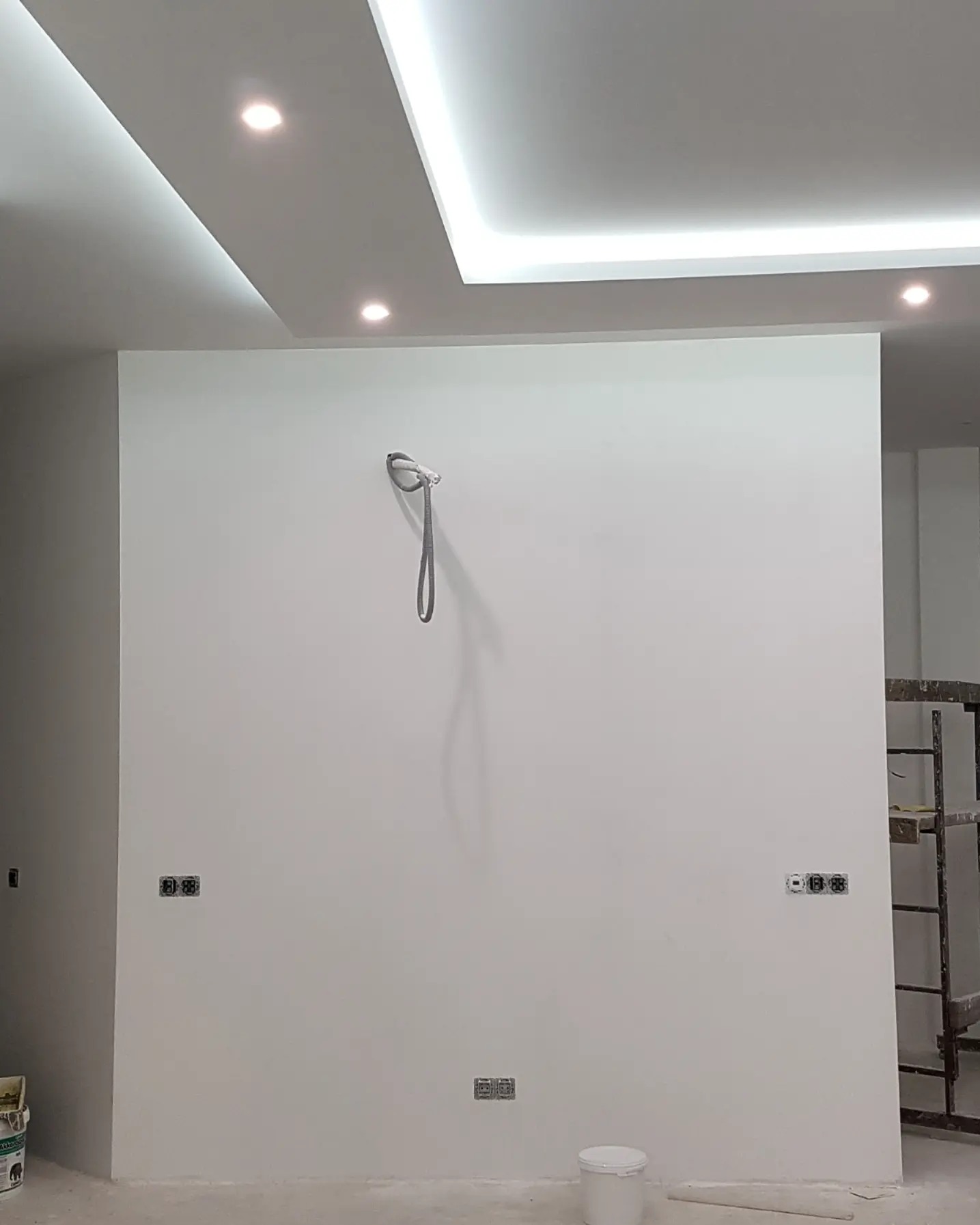 Świeżo pomalowana na biało ściana z widocznymi gniazdkami elektrycznymi i wystającym kablem, sufit z nowoczesnym oświetleniem LED, drabina malarska w tle.