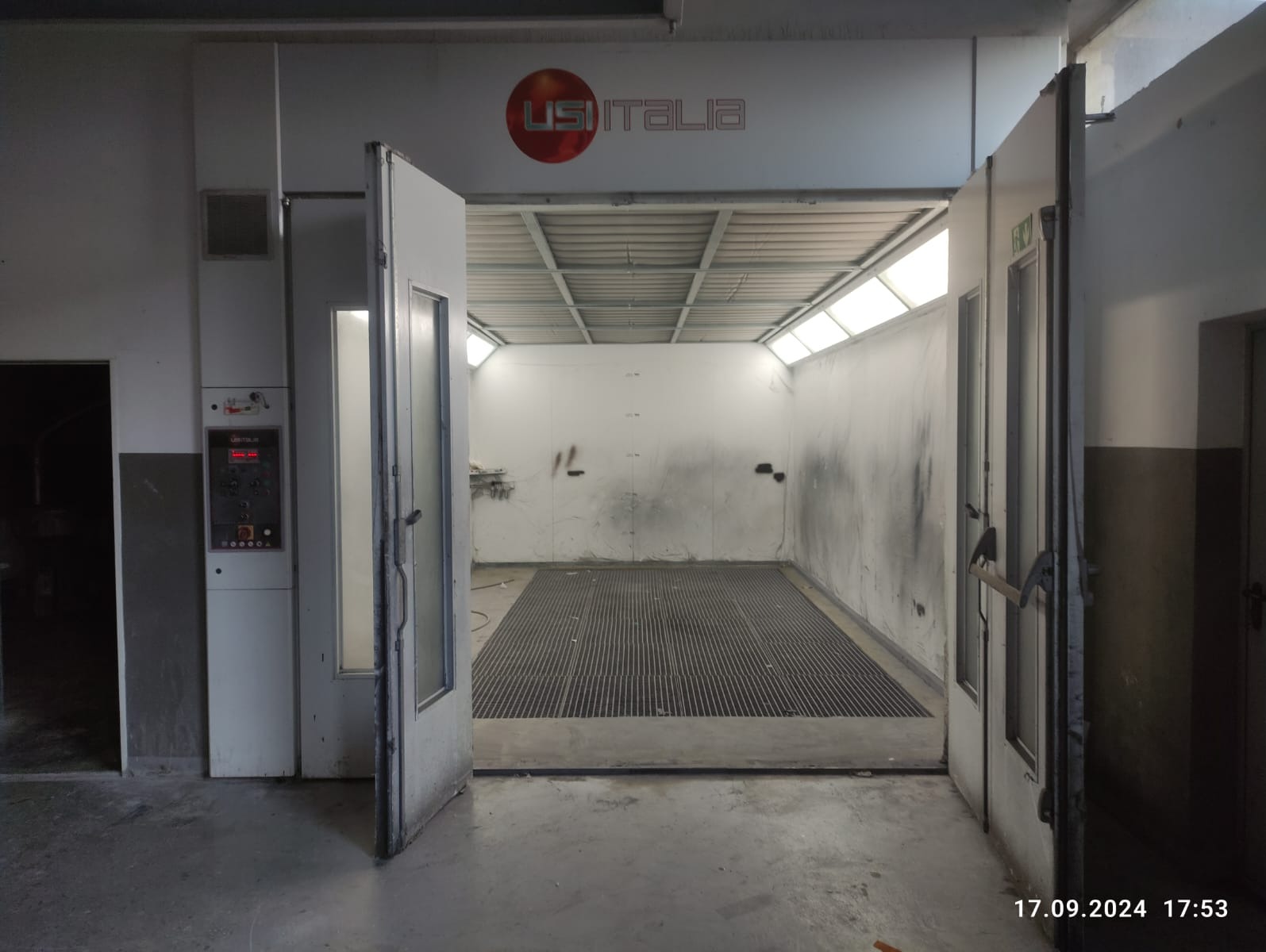 Pusta komora lakiernicza z otwartymi drzwiami, widoczne ślady użytkowania na ścianach i podłodze z kratką, logo USI ITALIA nad wejściem.