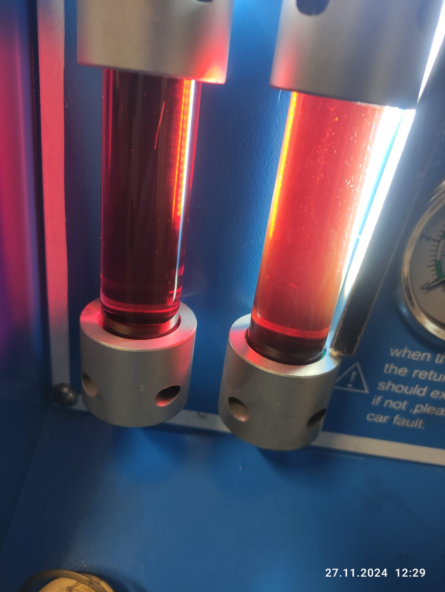Dwa przezroczyste cylindry z czerwoną cieczą w srebrnych mocowaniach na niebieskim urządzeniu diagnostycznym, prawdopodobnie podczas wymiany płynu w automatycznej skrzyni biegów.