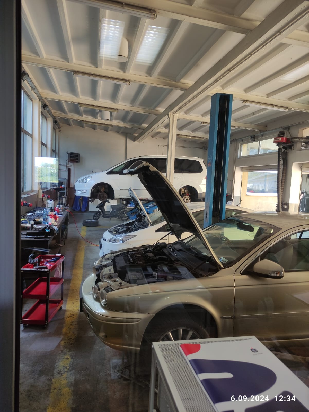 Wnętrze warsztatu samochodowego z trzema pojazdami: złoty Jaguar z otwartą maską na pierwszym planie, biały Ford z otwartą maską w tle oraz biały van na podnośniku, widok przez szybę.