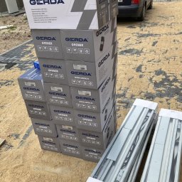 MAR-BUD Marcin Błaszczak - Stos szarych pudełek Gerda Opener z automatyką do bram ustawiony na piasku obok dwóch długich, zapakowanych szyn montażowych; w tle zaparkowany granatowy samochód osobowy i plac budowy.