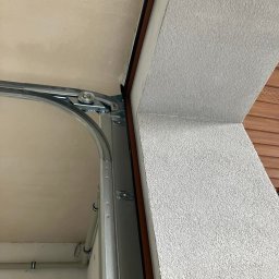 MAR-BUD Marcin Błaszczak - Wnętrze garażu z widocznym mechanizmem bramy segmentowej, szyny prowadzące z rolkami oraz fragment ściany z szorstkim tynkiem.