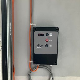 MAR-BUD Marcin Błaszczak - Panel sterowania bramy garażowej Nice D-Pro zamontowany na ścianie w Kodniu, z widocznymi przewodami i fragmentem prowadnicy bramy.