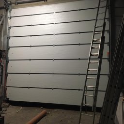 MAR-BUD Marcin Błaszczak - Zamknięta, segmentowa brama garażowa w odcieniach szarości z widocznymi prowadnicami i sprężynami, z dwoma rozstawionymi drabinami aluminiowymi na pierwszym planie w surowym, niedoświetlonym...