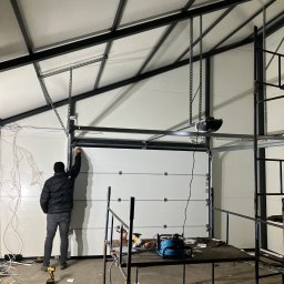 MAR-BUD Marcin Błaszczak - Instalacja segmentowej bramy garażowej: mężczyzna w kurtce montuje prowadnicę na tle surowego wnętrza hali, widoczny automat do bramy, narzędzia i rusztowanie.