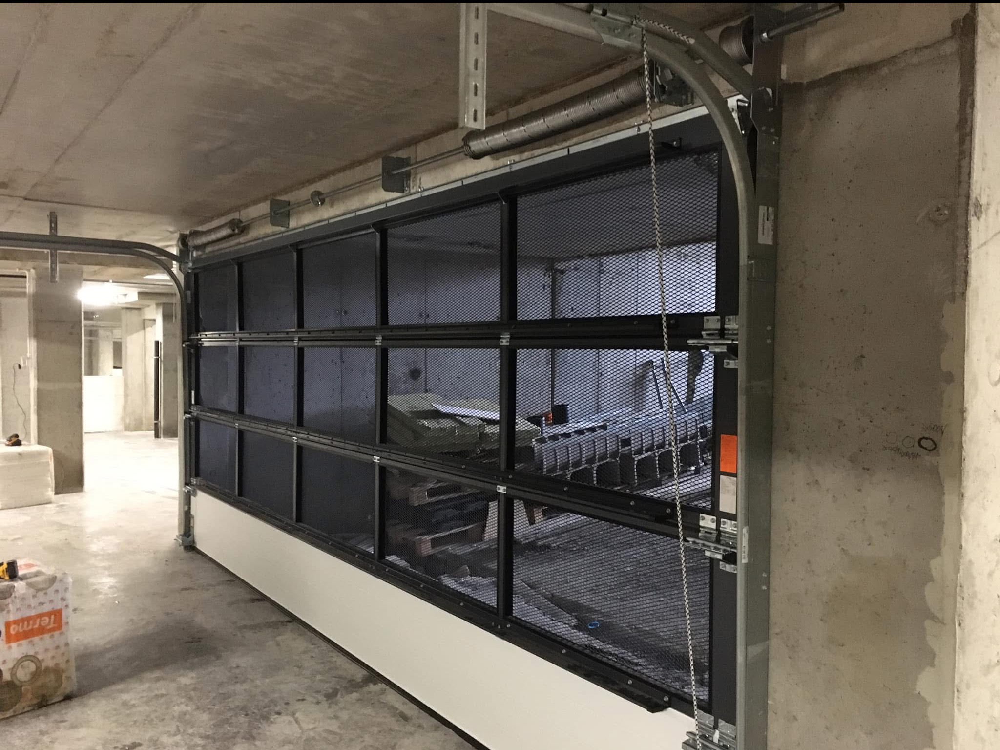 Metalowa brama garażowa segmentowa z siatką, widoczna od wewnątrz garażu, w trakcie montażu, z widocznymi elementami konstrukcyjnymi i opakowaniem materiałów budowlanych w tle.