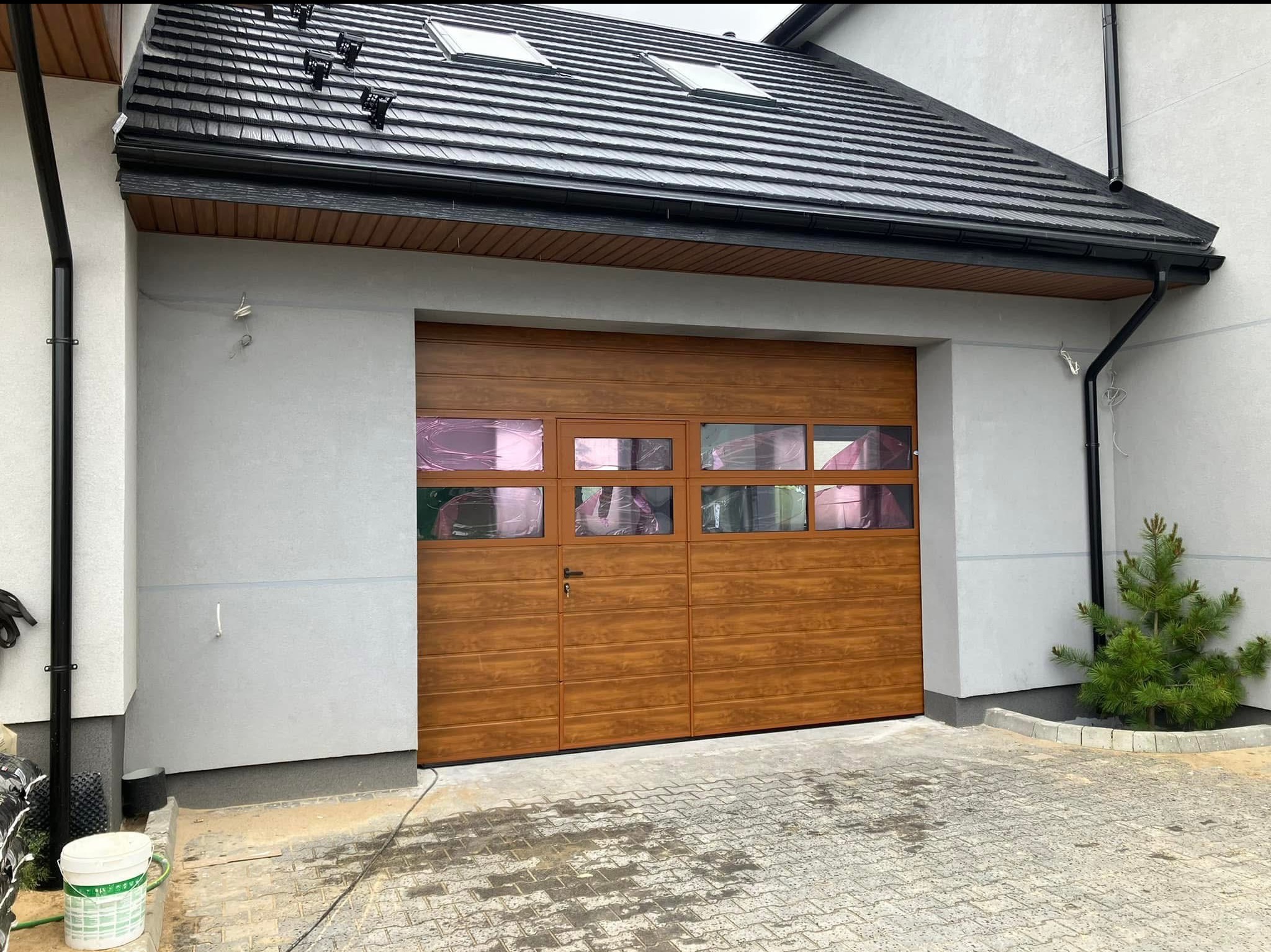 Segmentowa brama garażowa w kolorze drewna, z widocznymi szybami zabezpieczonymi folią ochronną, zamontowana w jasnoszarym garażu z czarnym dachem i rynnami, przed garażem kostka brukowa i mała...