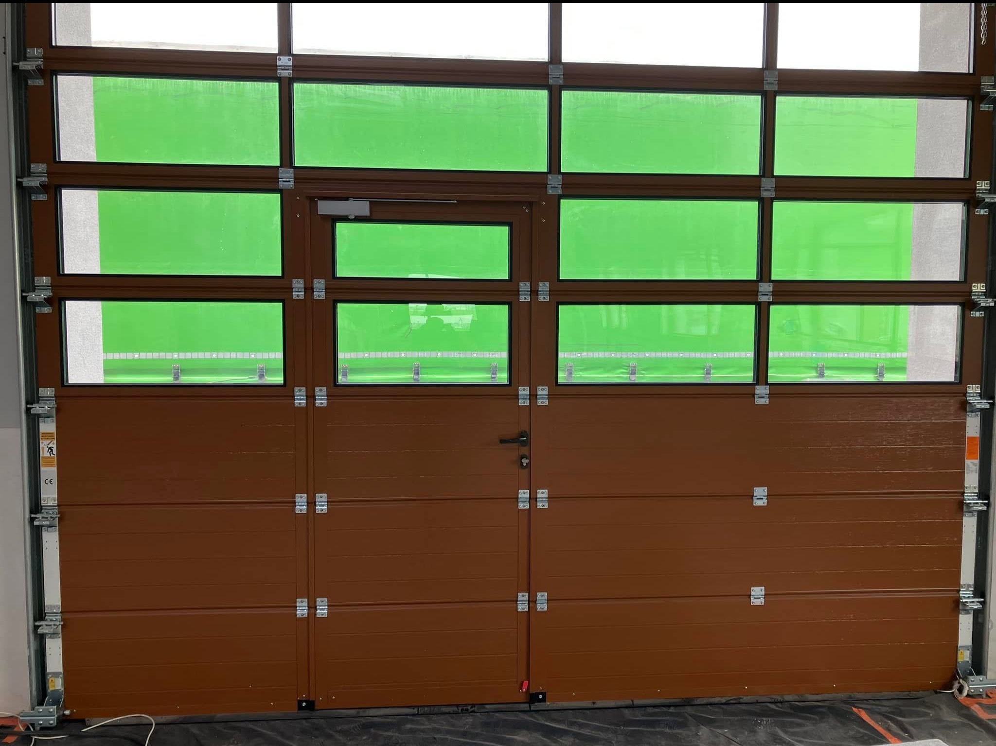 Segmentowa brama garażowa w kolorze brązowym z przeszklonymi panelami w górnej części, widoczne elementy montażowe i zielone tło za szybami.