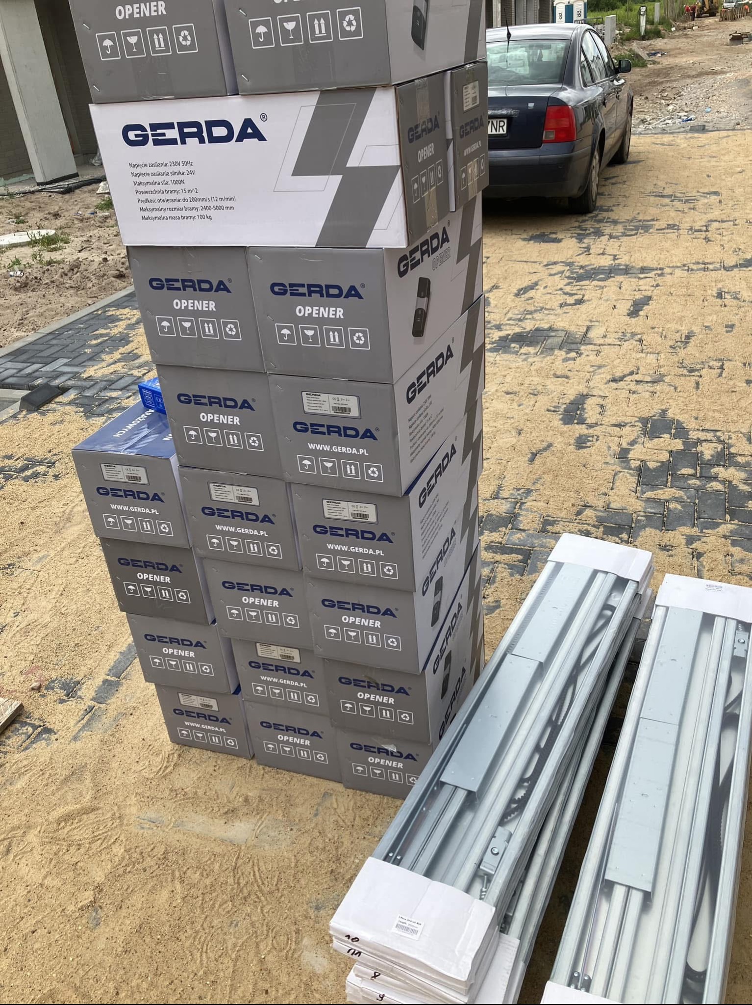 Stos szarych pudełek Gerda Opener z automatyką do bram ustawiony na piasku obok dwóch długich, zapakowanych szyn montażowych; w tle zaparkowany granatowy samochód osobowy i plac budowy.