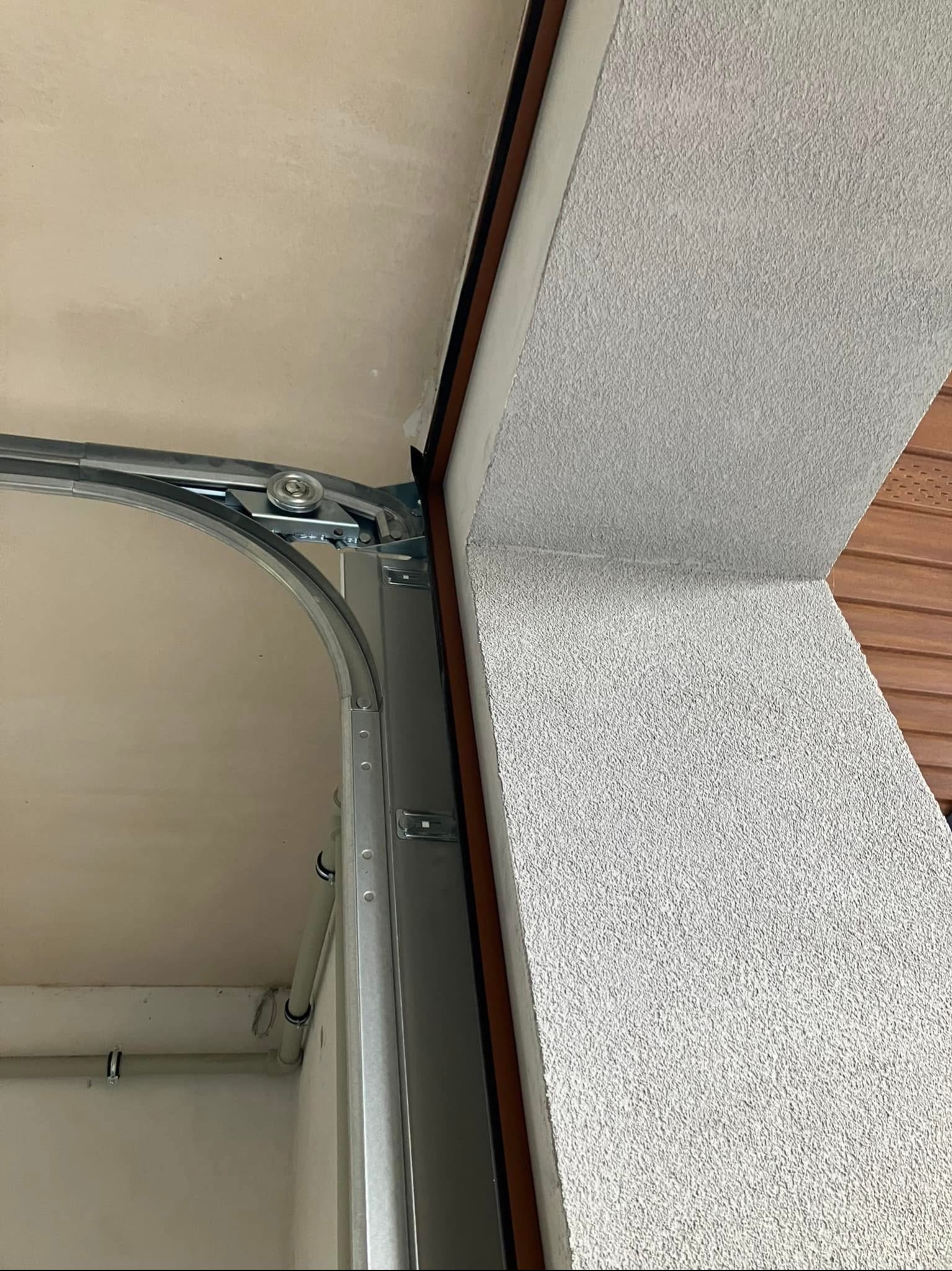 Wnętrze garażu z widocznym mechanizmem bramy segmentowej, szyny prowadzące z rolkami oraz fragment ściany z szorstkim tynkiem.