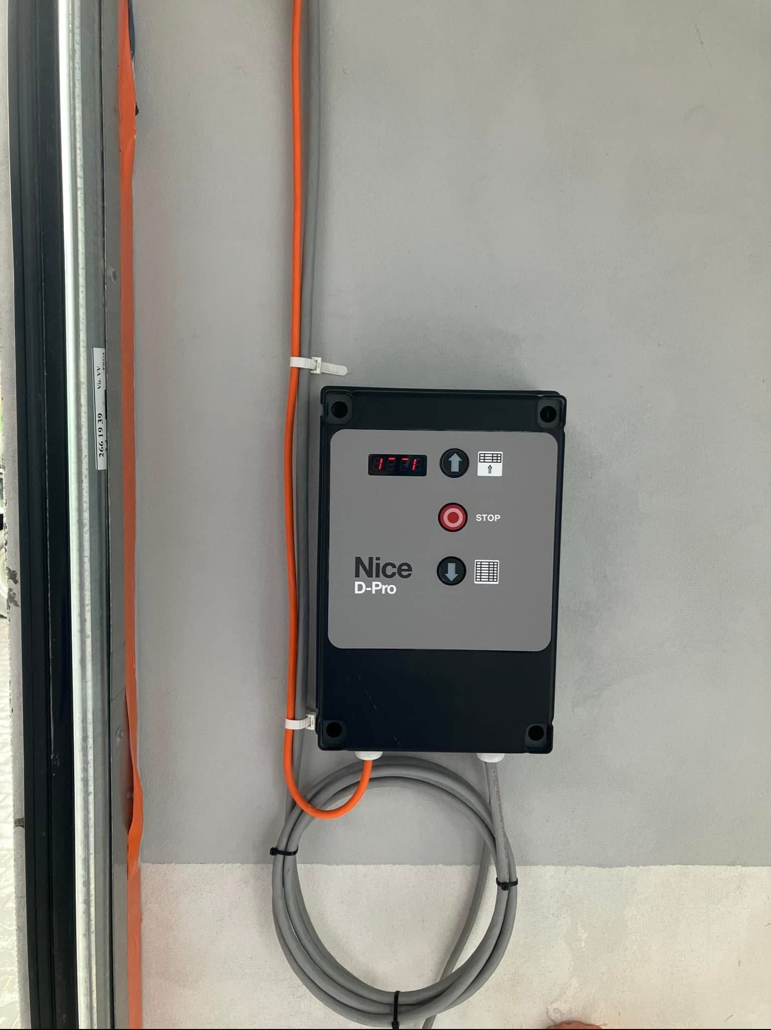 Panel sterowania bramy garażowej Nice D-Pro zamontowany na ścianie w Kodniu, z widocznymi przewodami i fragmentem prowadnicy bramy.