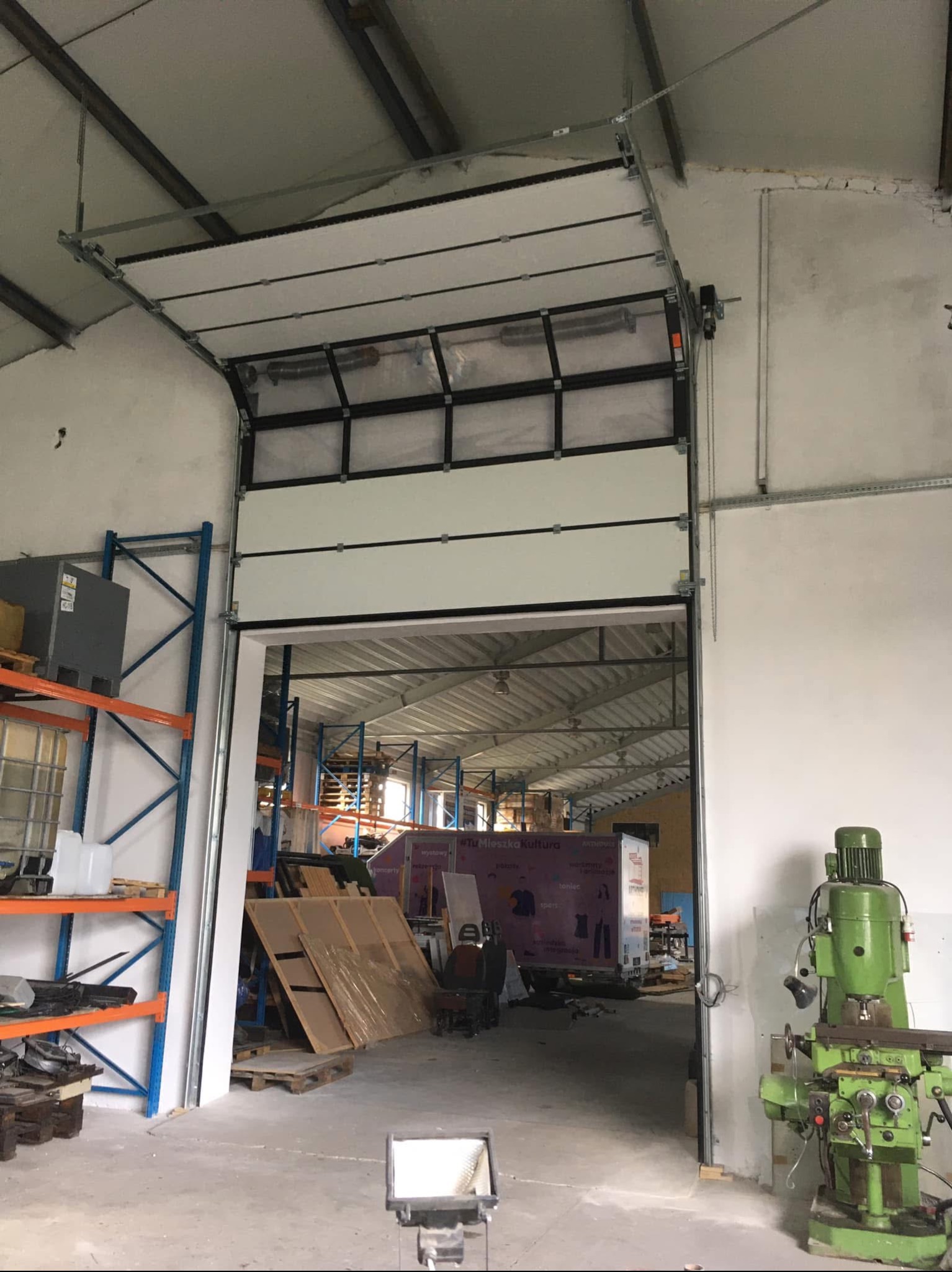 Częściowo otwarta segmentowa brama garażowa w hali magazynowej, widoczne prowadnice i mechanizm podnoszenia, w tle regały i maszyny.