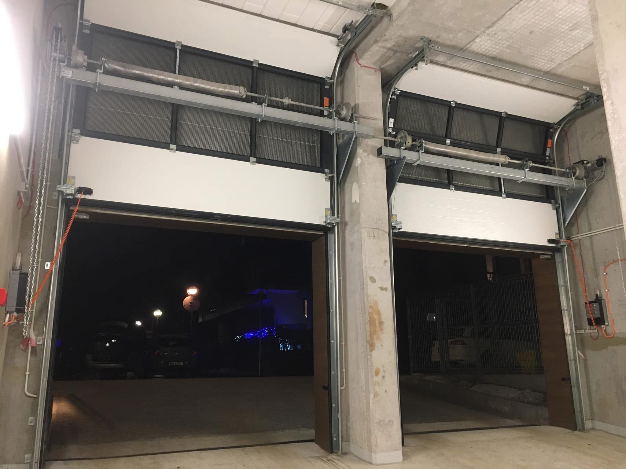 Dwie otwarte, segmentowe bramy garażowe w hali garażowej. Widoczne mechanizmy sprężynowe i prowadnice, a w tle nocny widok na oświetlony dom i zaparkowane samochody.