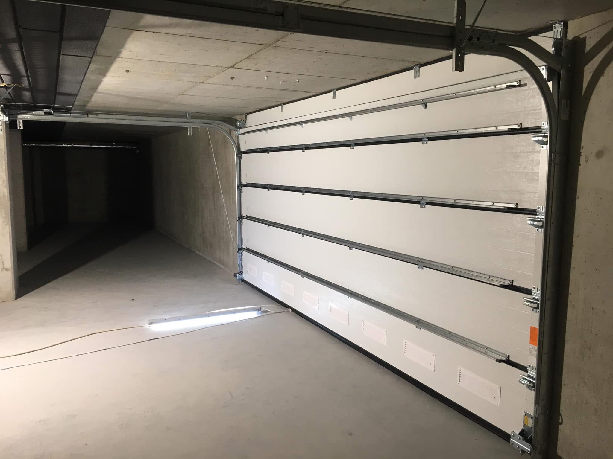 Częściowo otwarta, segmentowa brama garażowa w betonowym garażu, widoczne prowadnice i oświetlenie tymczasowe na podłodze.