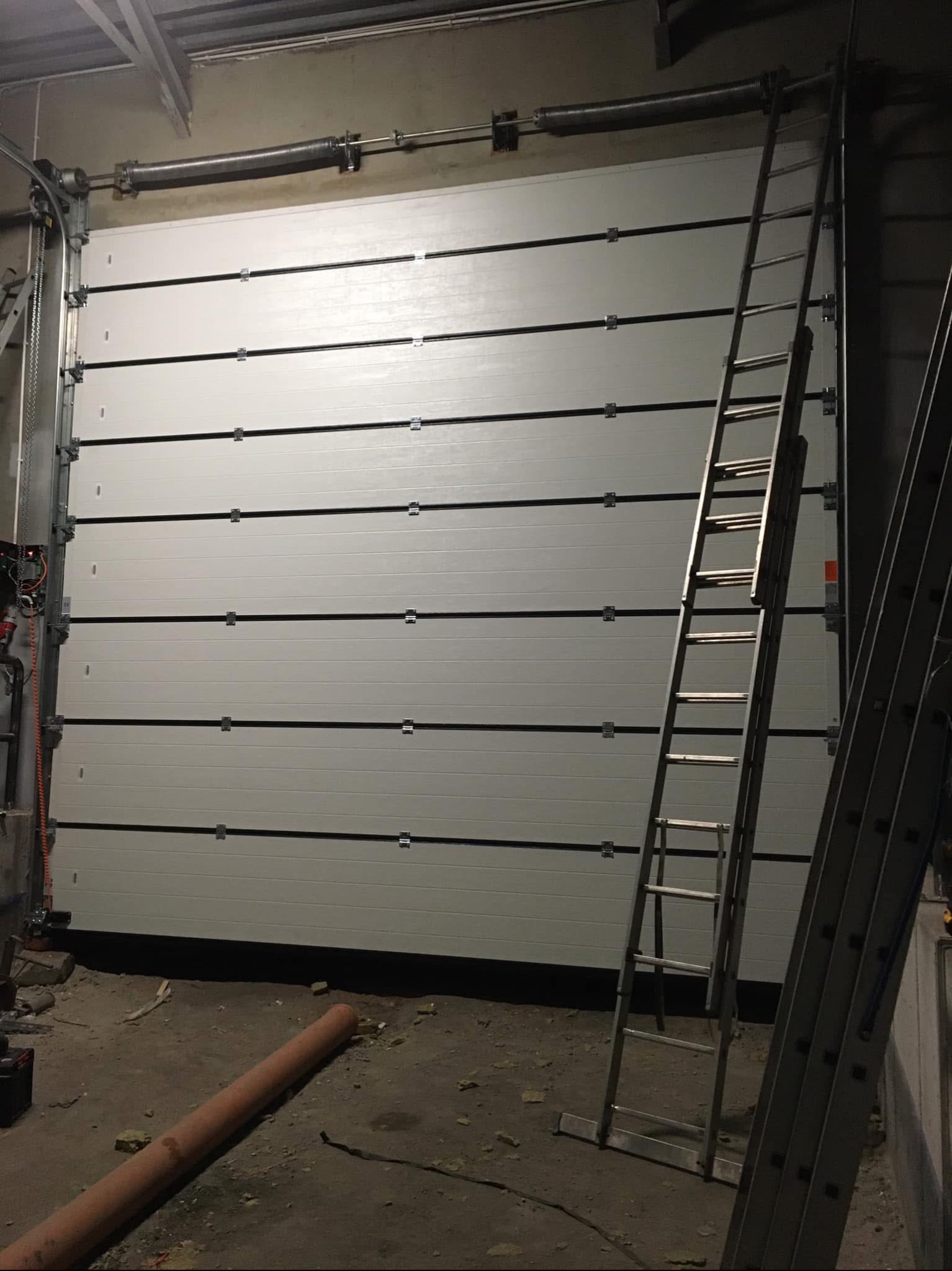 Zamknięta, segmentowa brama garażowa w odcieniach szarości z widocznymi prowadnicami i sprężynami, z dwoma rozstawionymi drabinami aluminiowymi na pierwszym planie w surowym, niedoświetlonym...