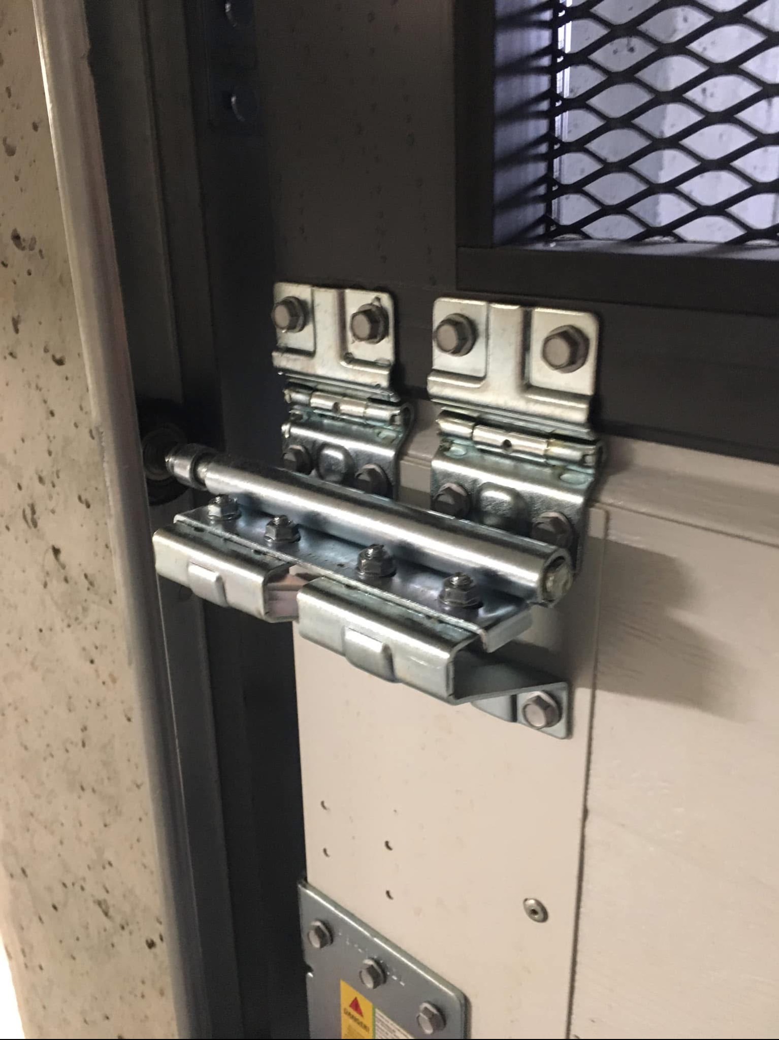 Szczegół metalowych zawiasów i mechanizmu blokującego na bramie garażowej z siatką w tle, widoczne śruby i elementy mocujące.