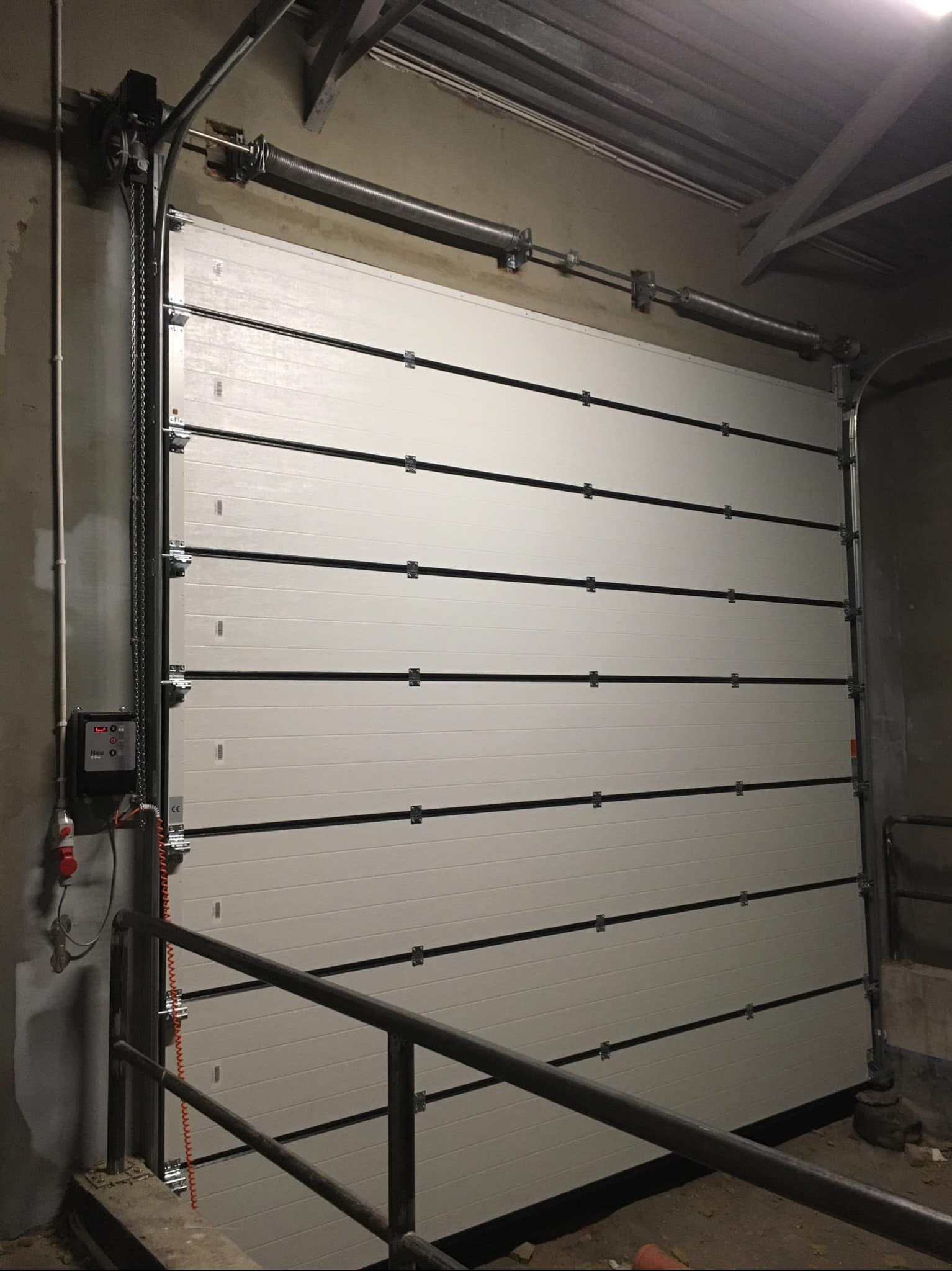 Zamknięta, segmentowa brama garażowa z automatyką i łańcuchem, widoczne sprężyny i sterownik z kablem spiralnym, zamontowana w surowym, przemysłowym wnętrzu.