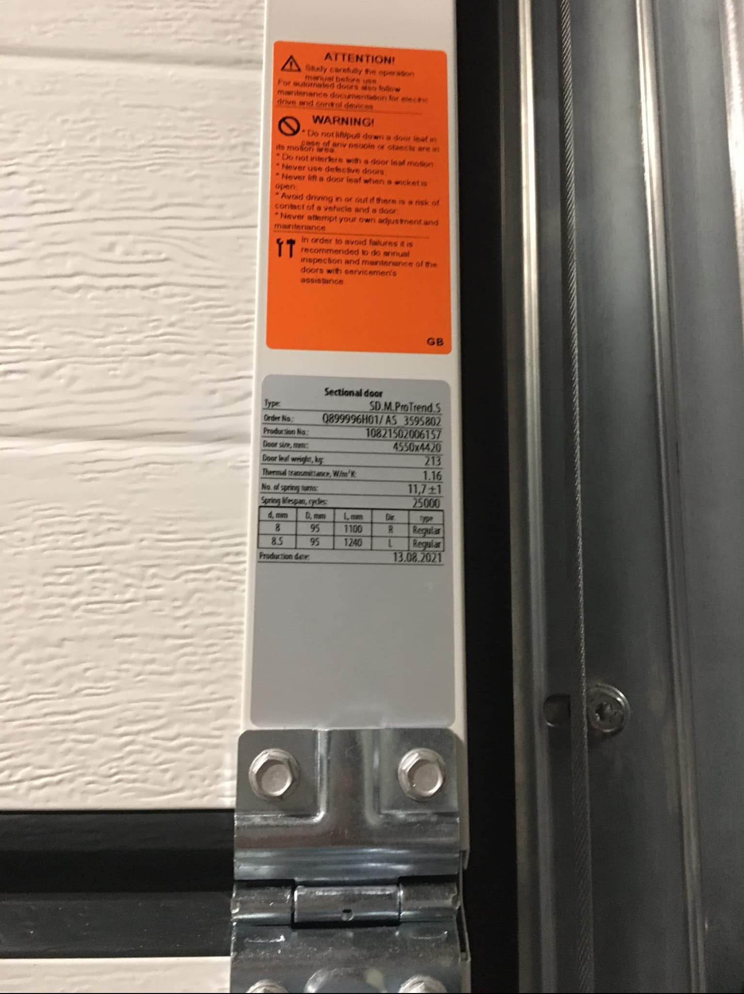 Szczegółowe zbliżenie na tabliczkę znamionową segmentowej bramy garażowej z informacjami technicznymi, ostrzeżeniami i datą produkcji 13.08.2021, widoczne śruby mocujące i fragment konstrukcji bramy.