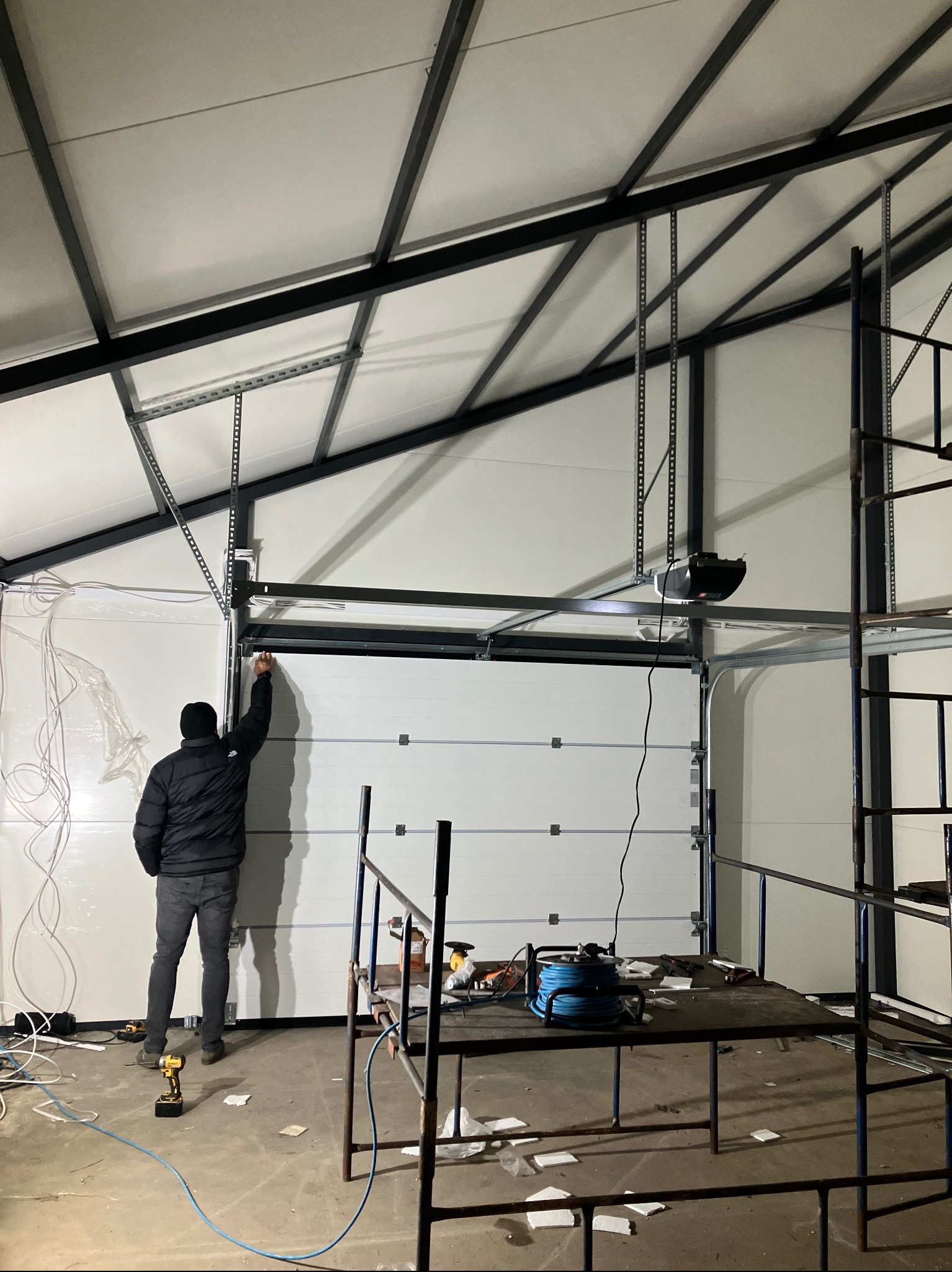 Instalacja segmentowej bramy garażowej: mężczyzna w kurtce montuje prowadnicę na tle surowego wnętrza hali, widoczny automat do bramy, narzędzia i rusztowanie.
