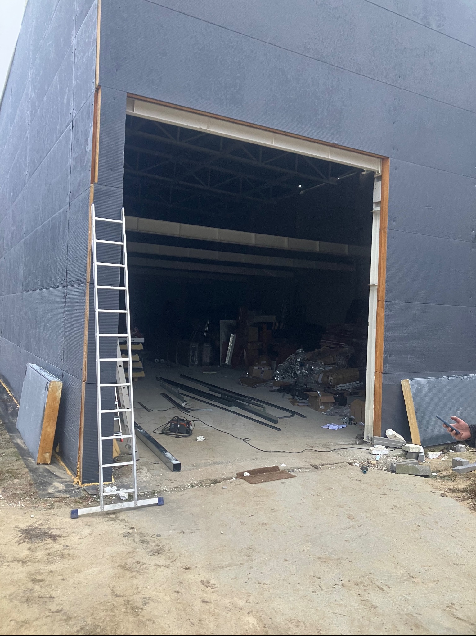Montaż bramy garażowej w hali produkcyjnej, widoczna drabina oparta o ścianę, elementy konstrukcyjne i narzędzia wewnątrz hali, surowy betonowy podjazd.