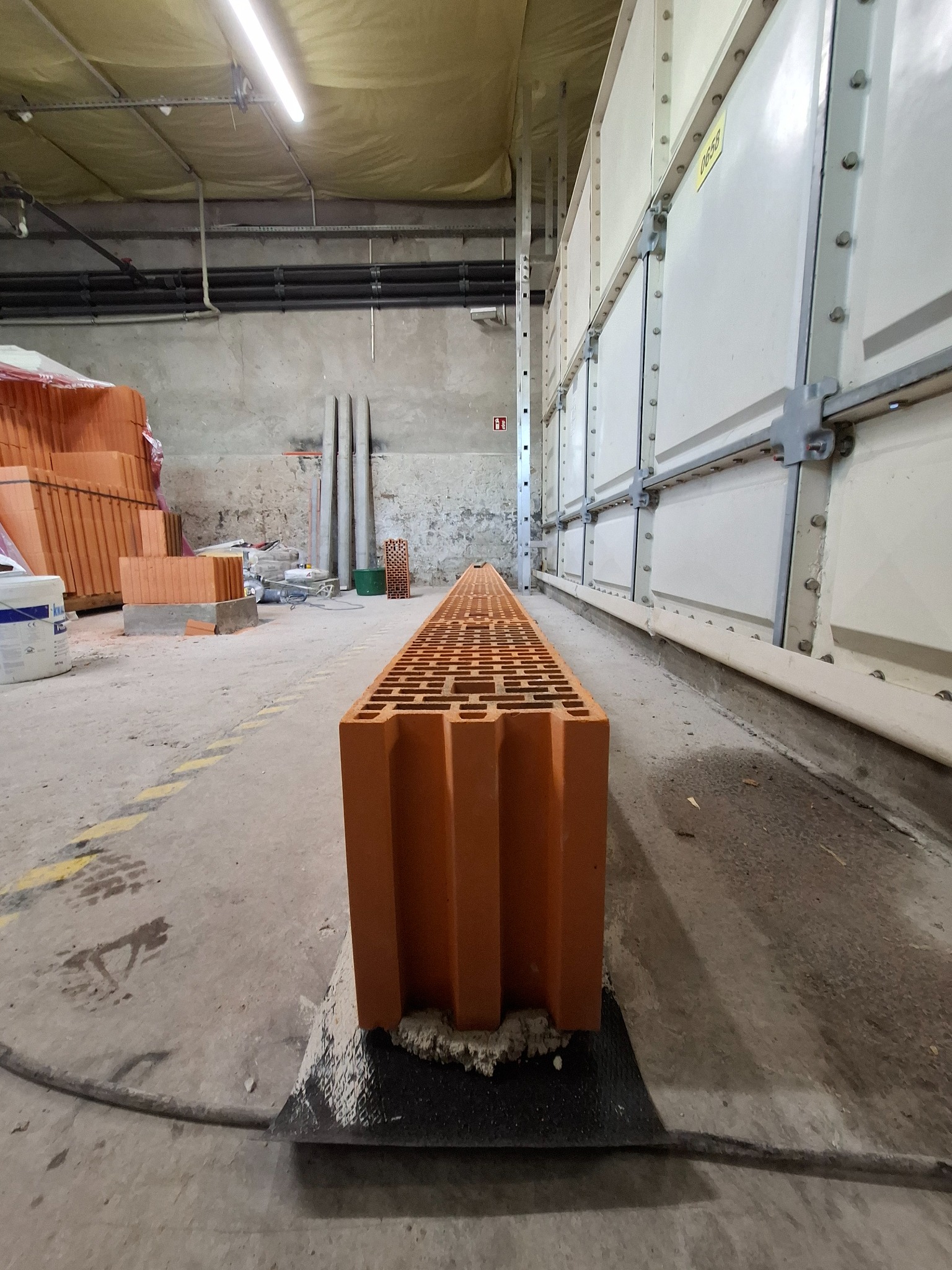 Długi, pustak ceramiczny ułożony na podłodze w hali produkcyjnej, widoczny z perspektywy podłogi, z zapasem cegieł w tle.