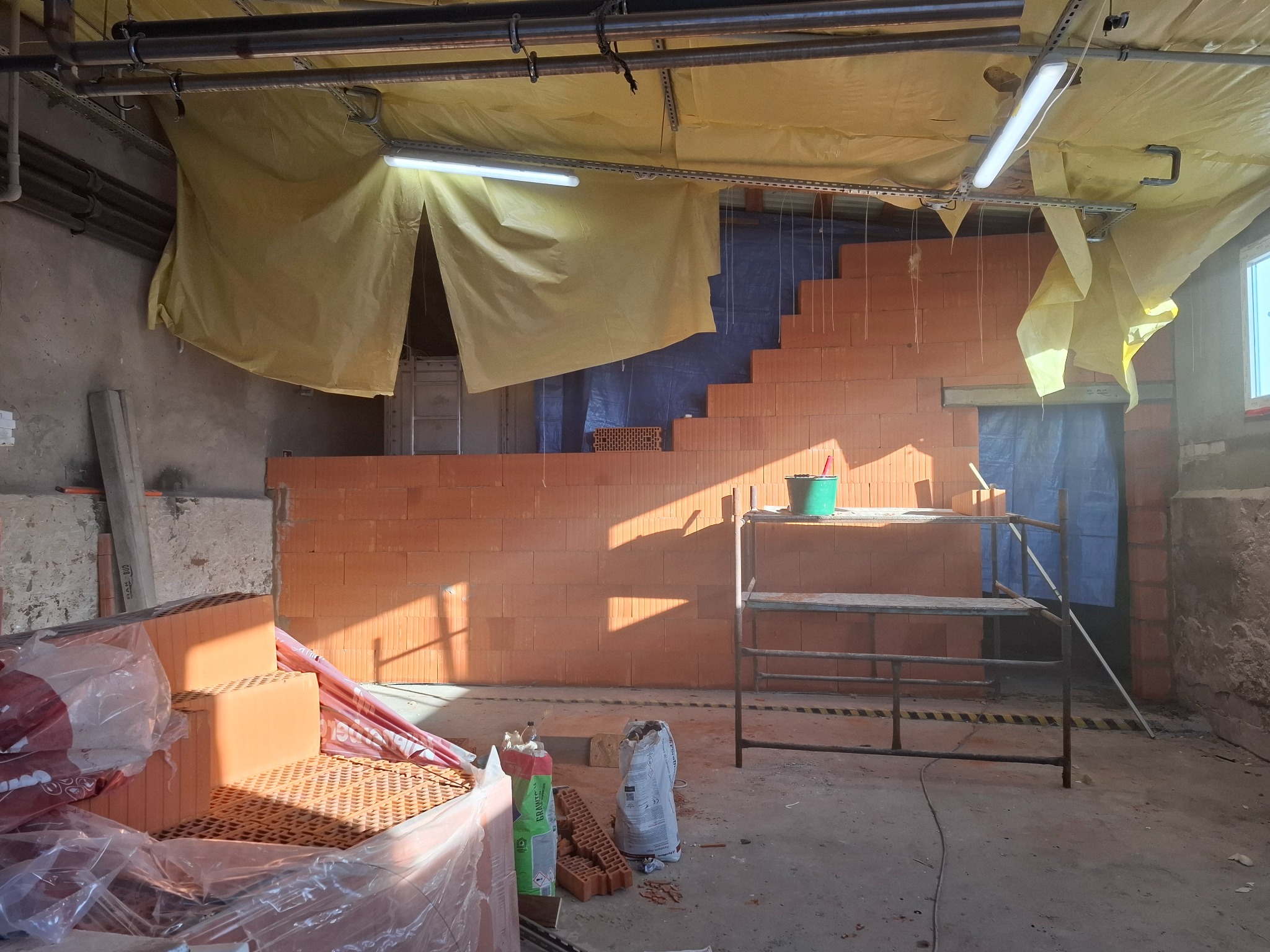 Wnętrze w trakcie murowania ścian z czerwonej cegły, widoczne rusztowanie, materiały budowlane i prowizoryczne oświetlenie pod żółtą folią zabezpieczającą.